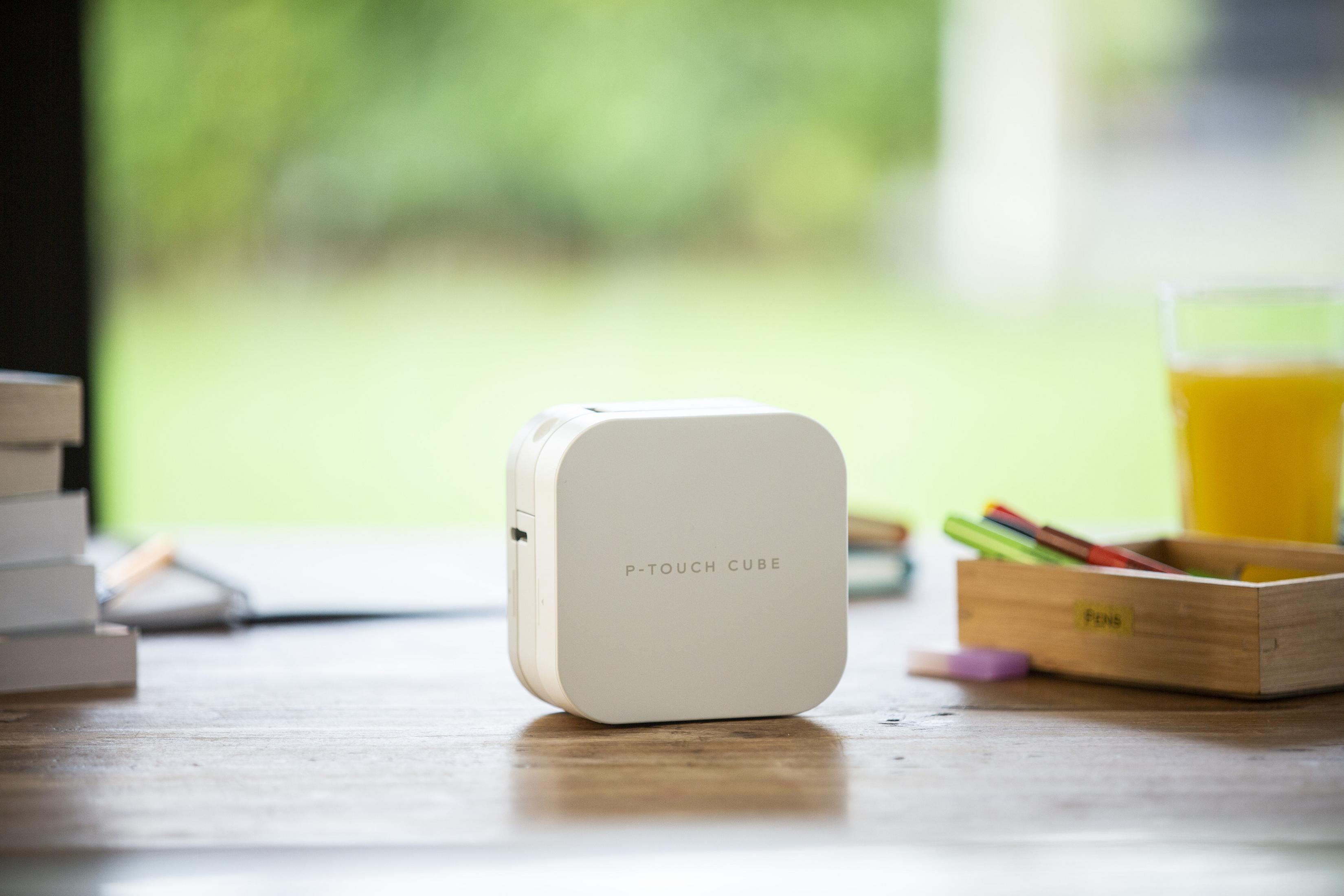 Étiqueteuse P-Touch Cube blanche sur un bureau avec des livres, des stylos et du jus d'orange.