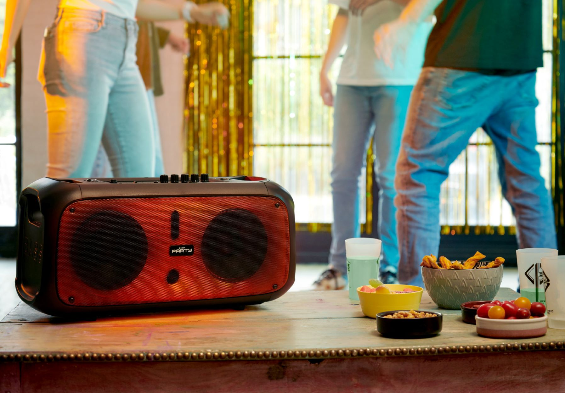 Przenośny głośnik na stole z ludźmi tańczącymi w tle. Widoczne przekąski.