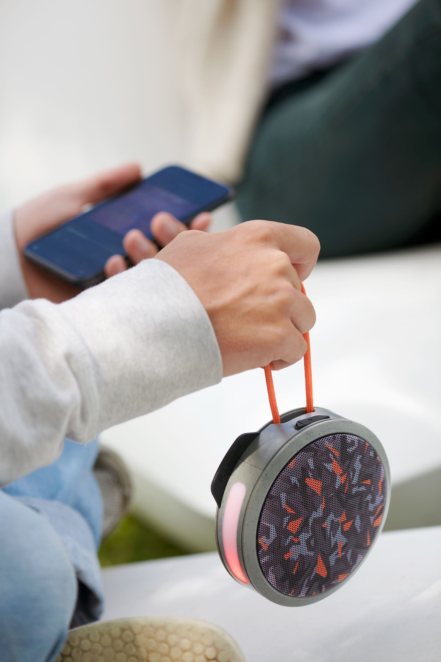 Una persona tiene in mano uno smartphone e un piccolo altoparlante rotondo con un cinturino arancione. L'altoparlante ha un motivo.