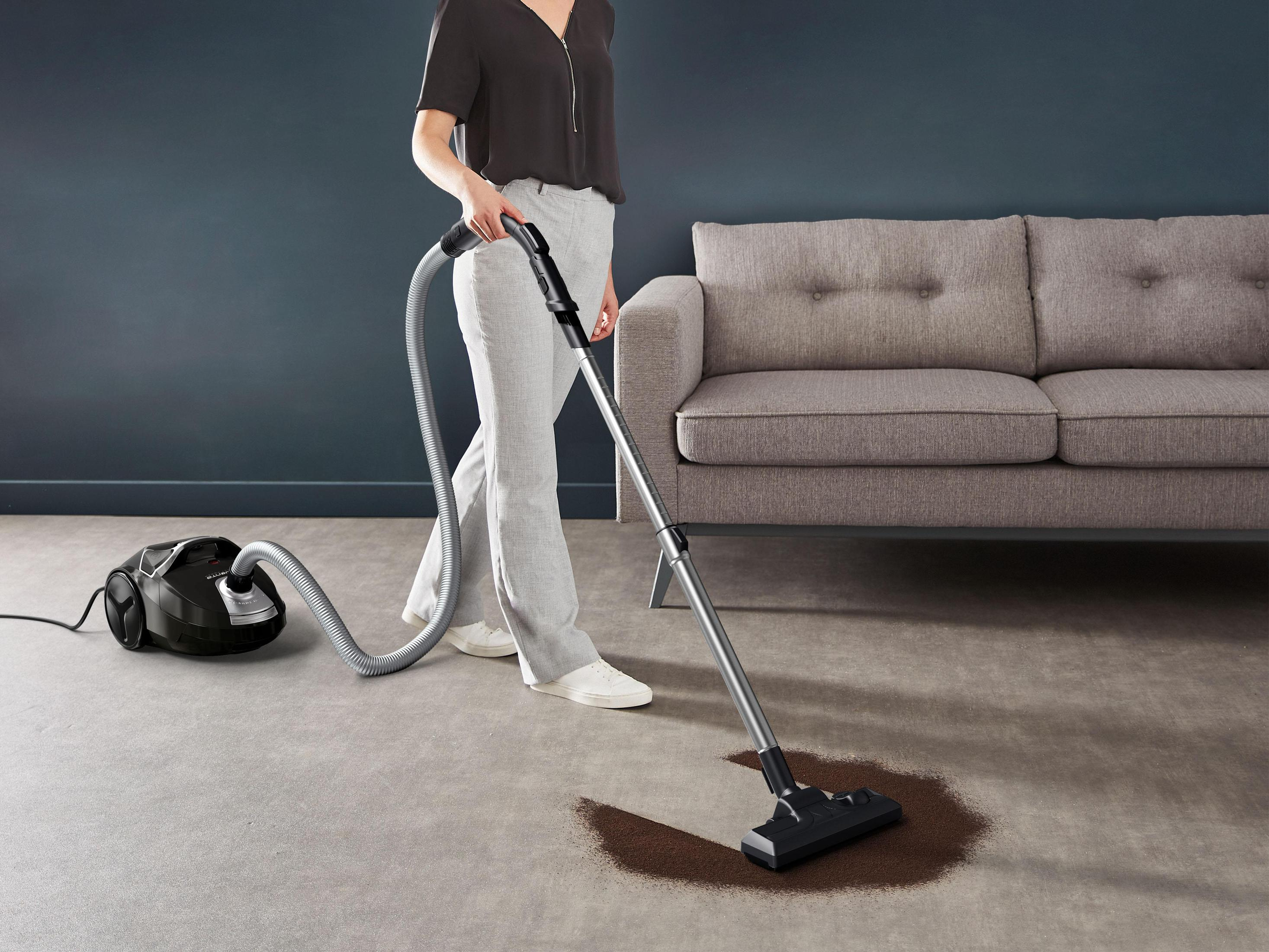 Une personne passe l'aspirateur sur un tapis, utilisant un aspirateur noir. Un canapé gris est en arrière-plan. Le tapis est gris.