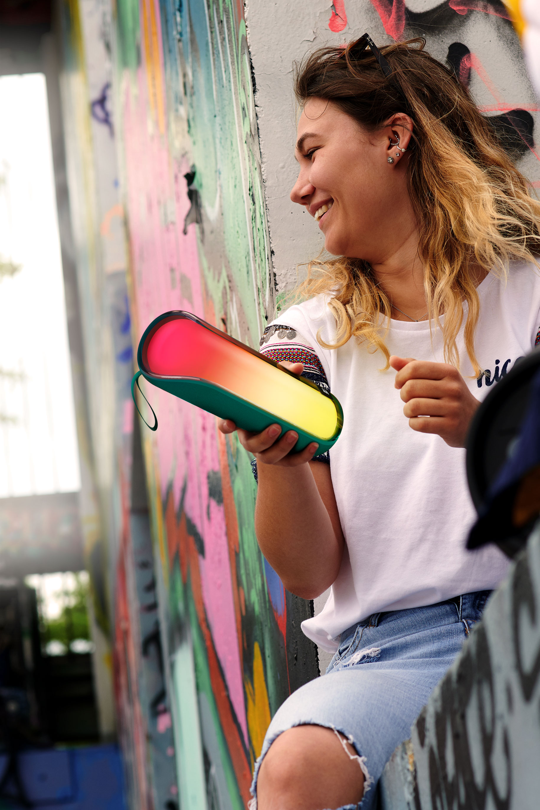 Kobieta uśmiecha się, trzymając kolorowy głośnik, siedząc przed ścianą z graffiti.