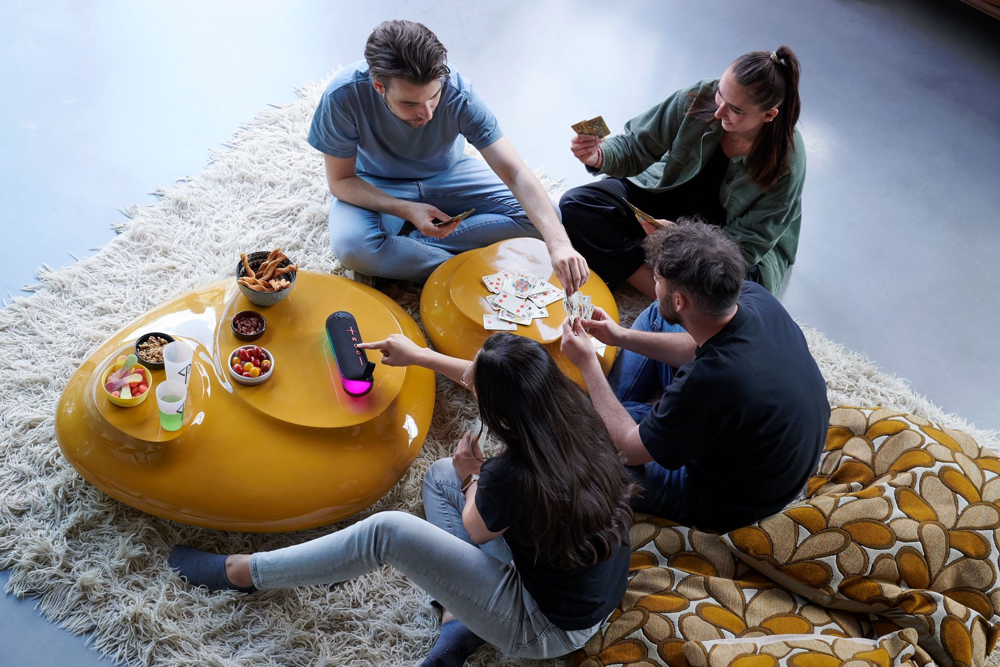 Cuatro personas jugando a las cartas en mesas amarillas. Aperitivos y bebidas en la mesa.