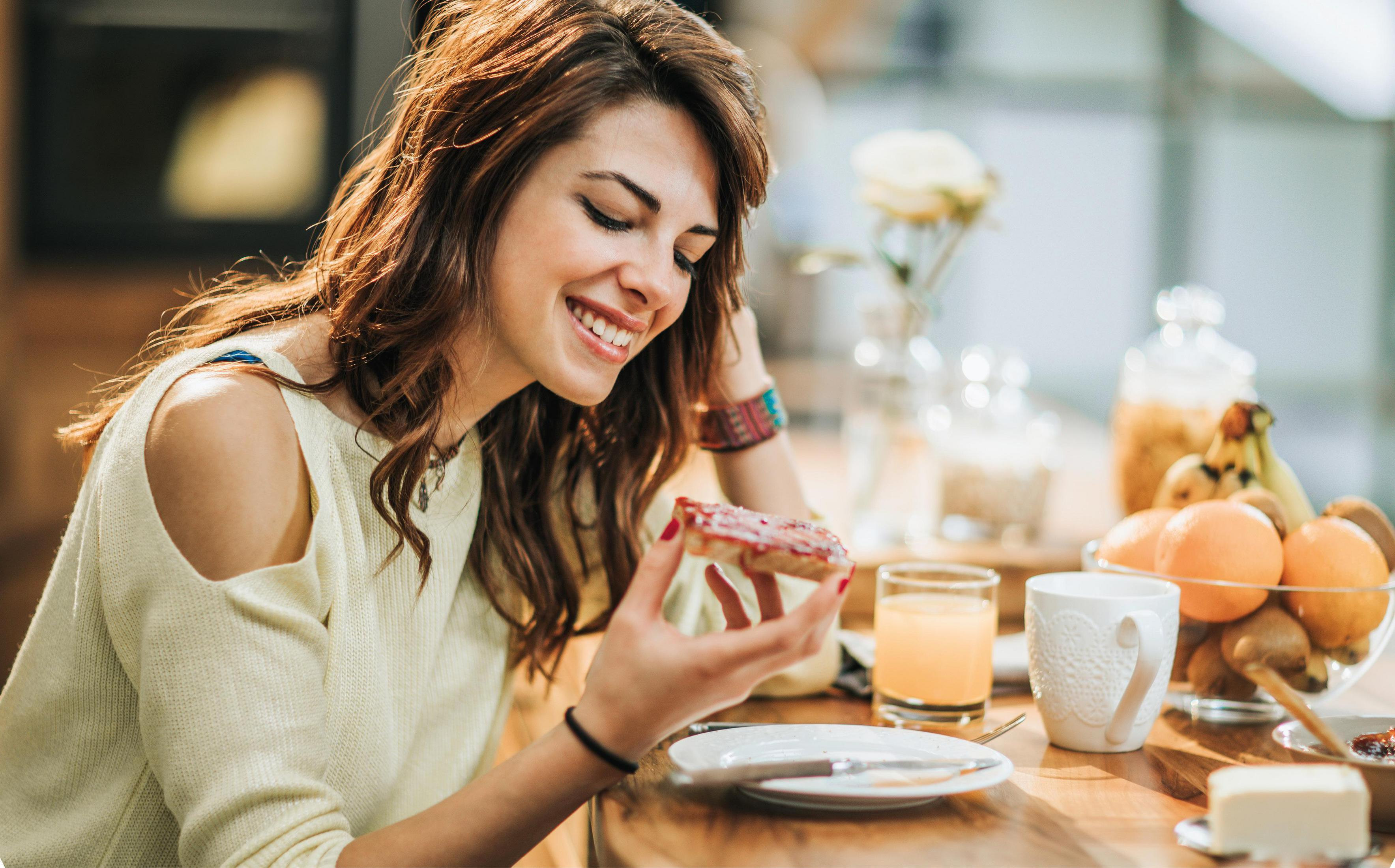 Una donna sorridente, che mangia un toast con marmellata, con frutta e caffè nelle vicinanze.