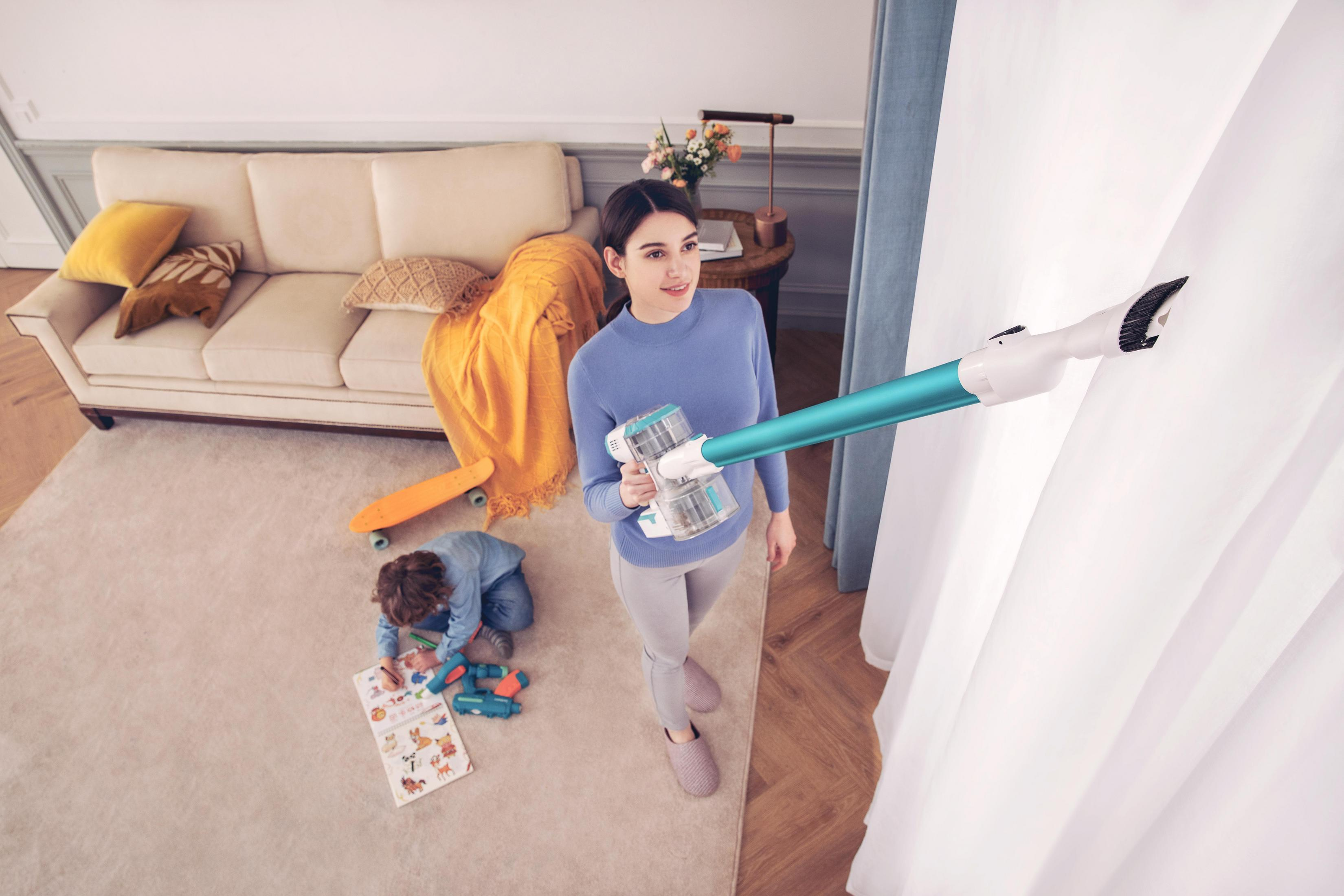 Femme aspirant un mur avec un aspirateur sans fil. Enfant jouant au sol, canapé et tapis en arrière-plan.