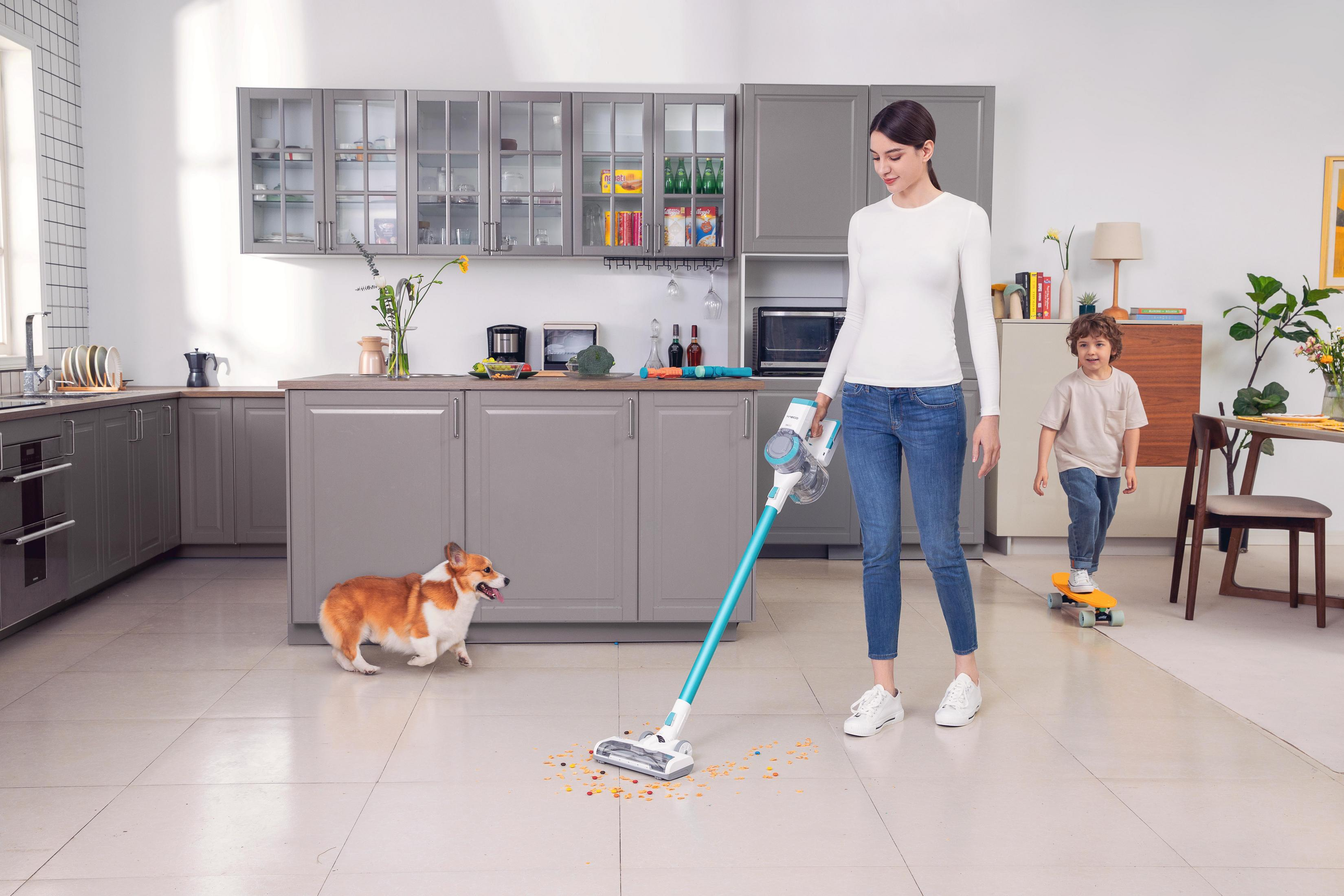 Une femme qui passe l'aspirateur dans une cuisine avec un enfant en skateboard et un chien.