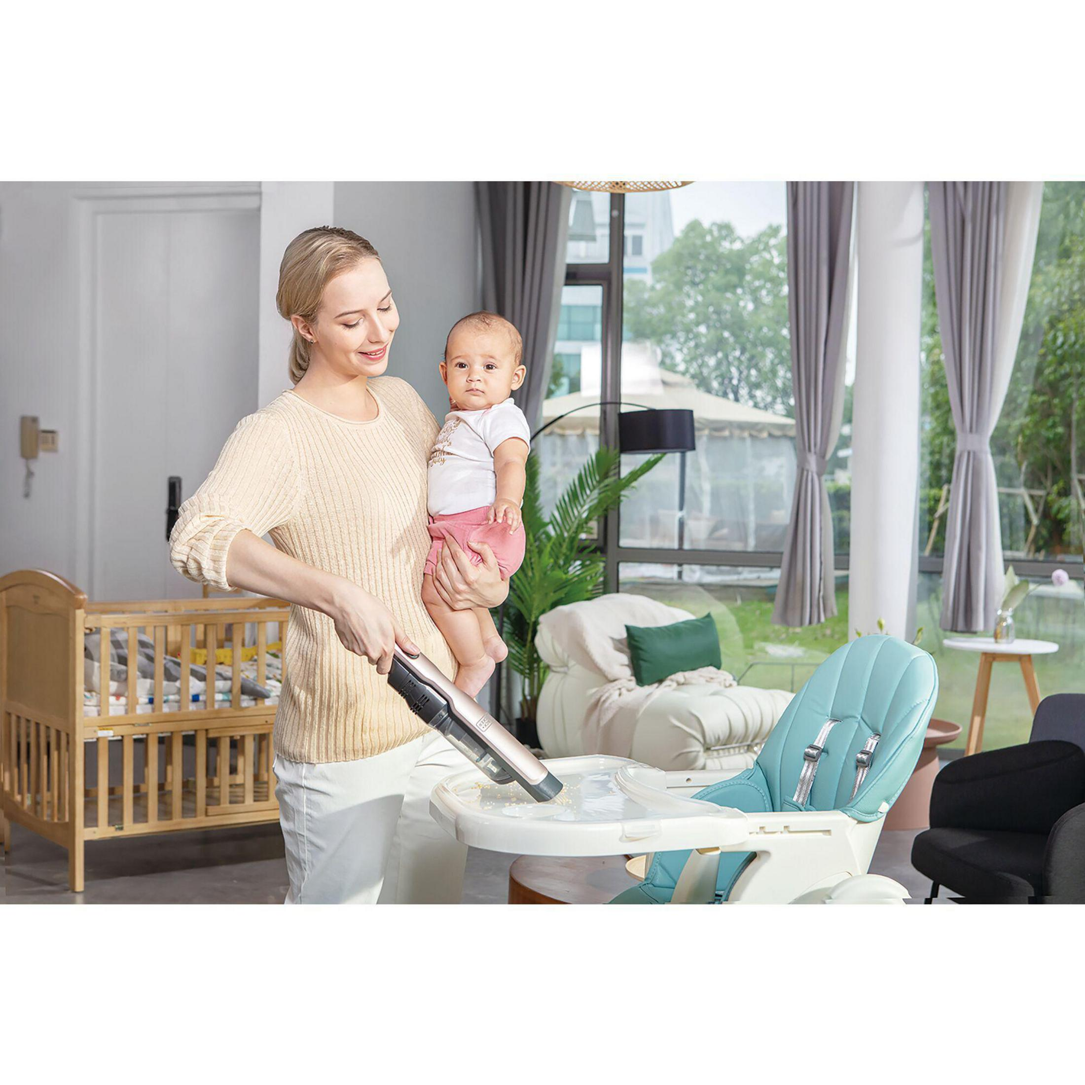 A woman holding a baby, using a vacuum cleaner on a high chair. A wooden crib is in the background.