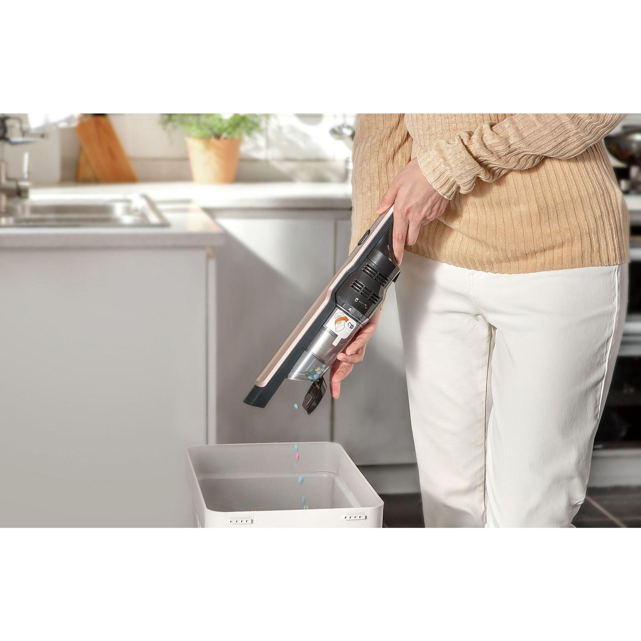 A person emptying a handheld vacuum cleaner into a white trash can in a kitchen setting.