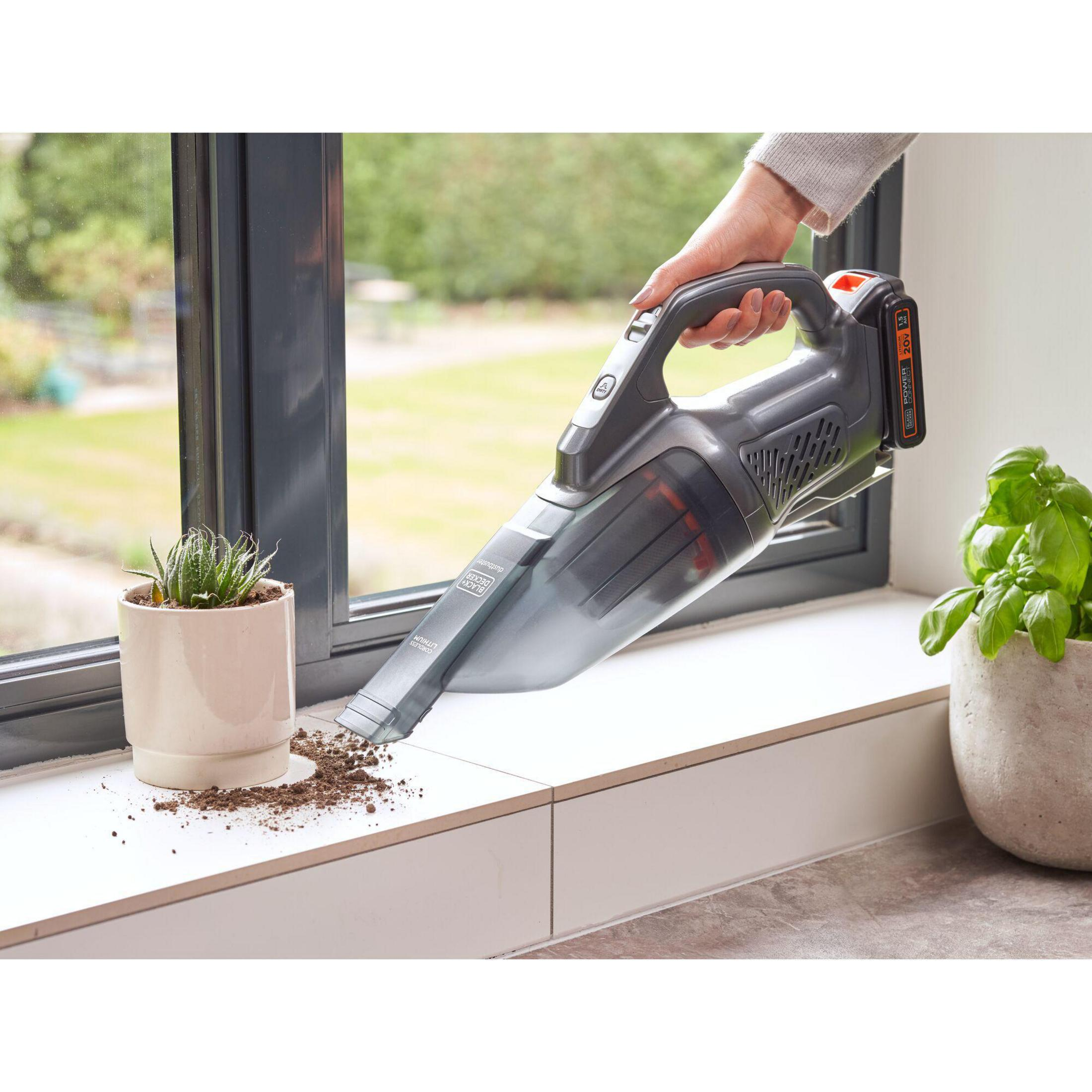 Eine Person reinigt mit einem Handstaubsauger ein Fensterbrett mit Schmutz und Pflanze.