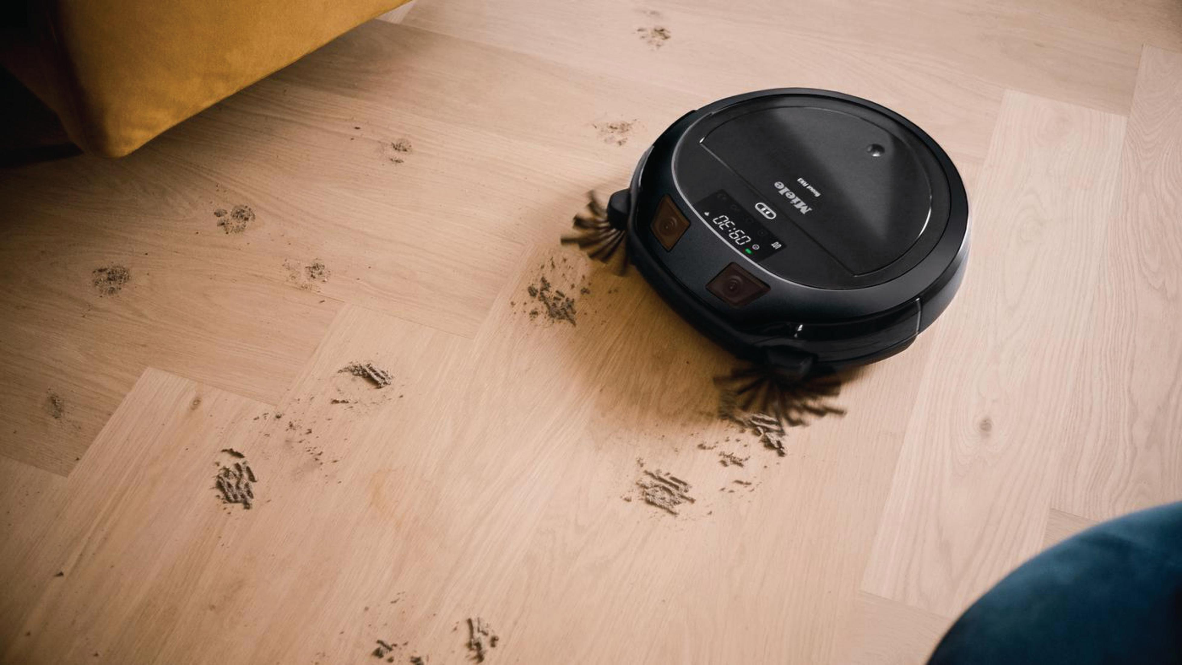 Roboter-Staubsauger auf Holzboden, reinigt. Staub und Schmutz sichtbar. Eine Ecke eines gelben Sofas sichtbar.