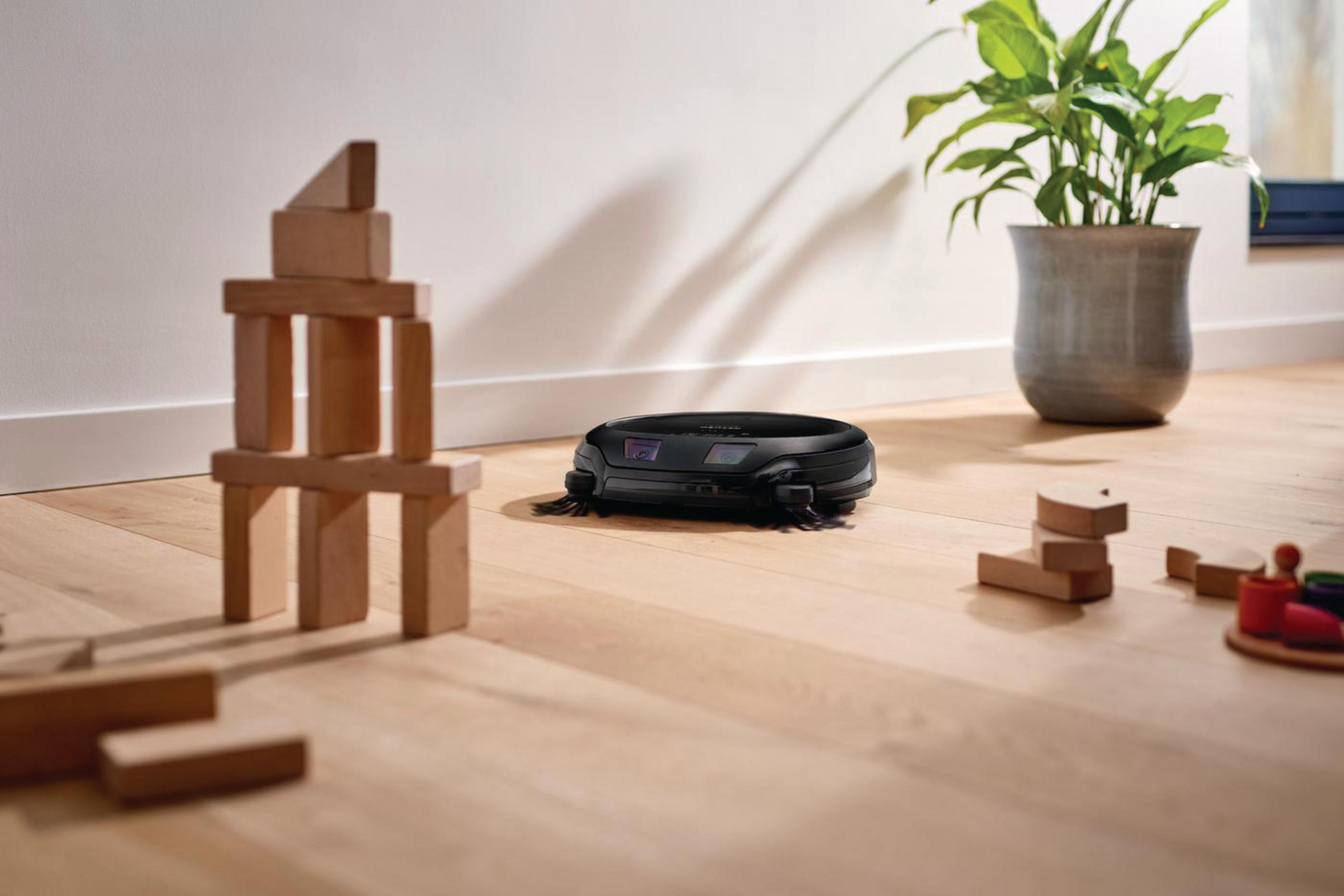 Ein schwarzer Roboter-Staubsauger in der Nähe von Holzblöcken und einer Pflanze auf Holzböden.