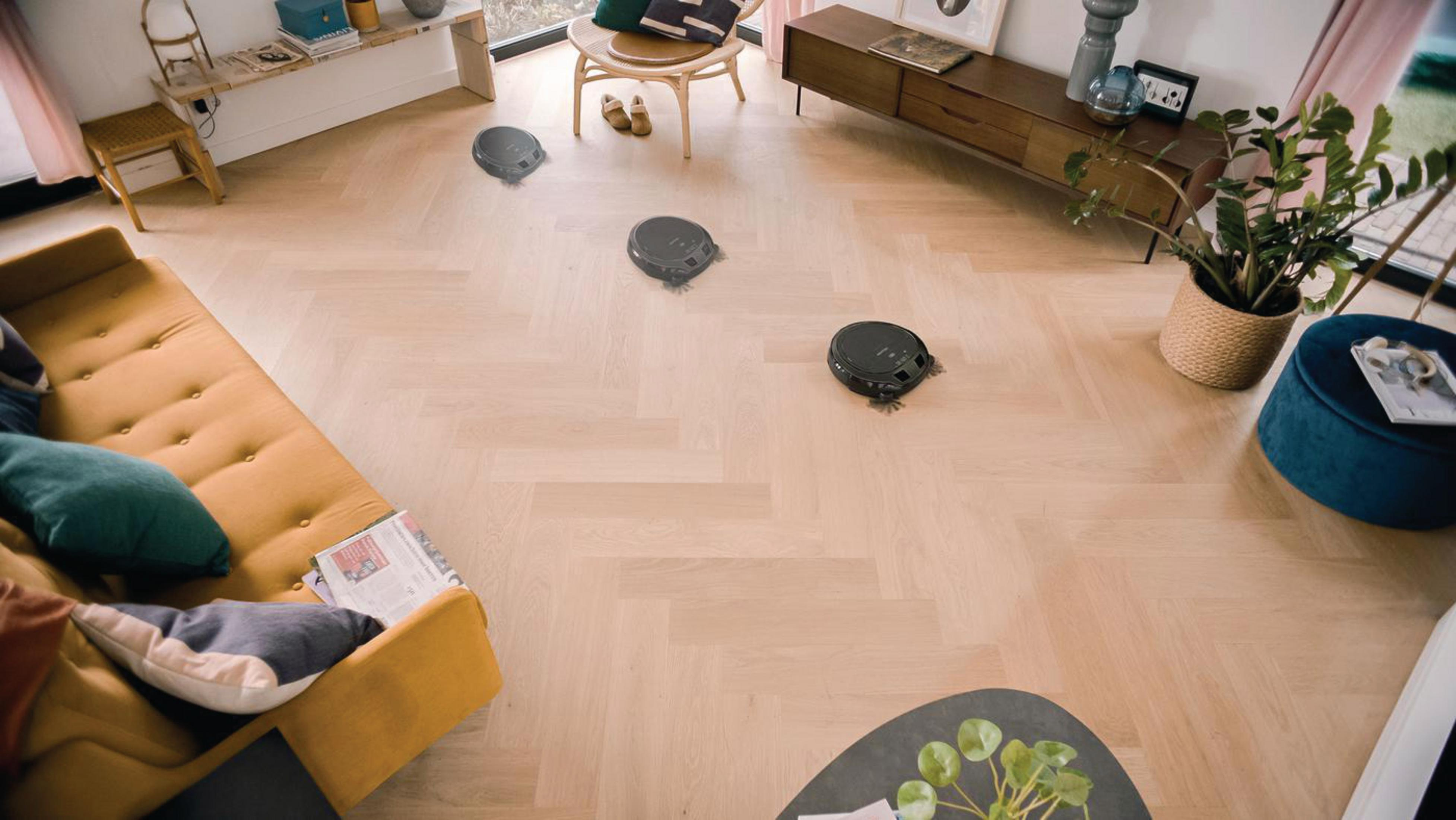 Draufsicht auf Raum mit drei Roboter-Staubsaugern, gelbem Sofa und Dekoration.