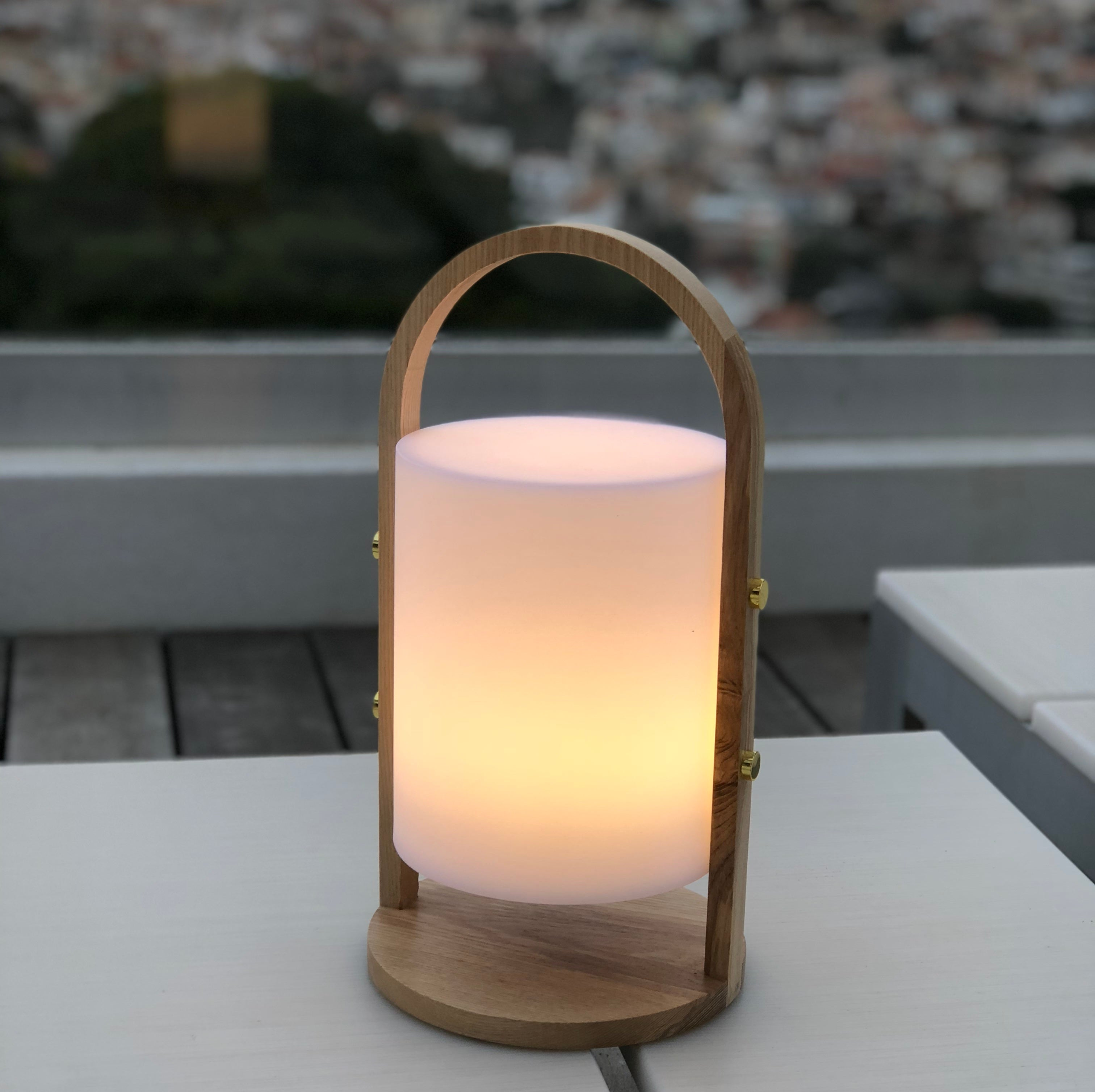 Holzlampe mit weißem Schein. Sie hat einen Griff und steht auf einem Tisch mit Stadtansicht im Hintergrund.