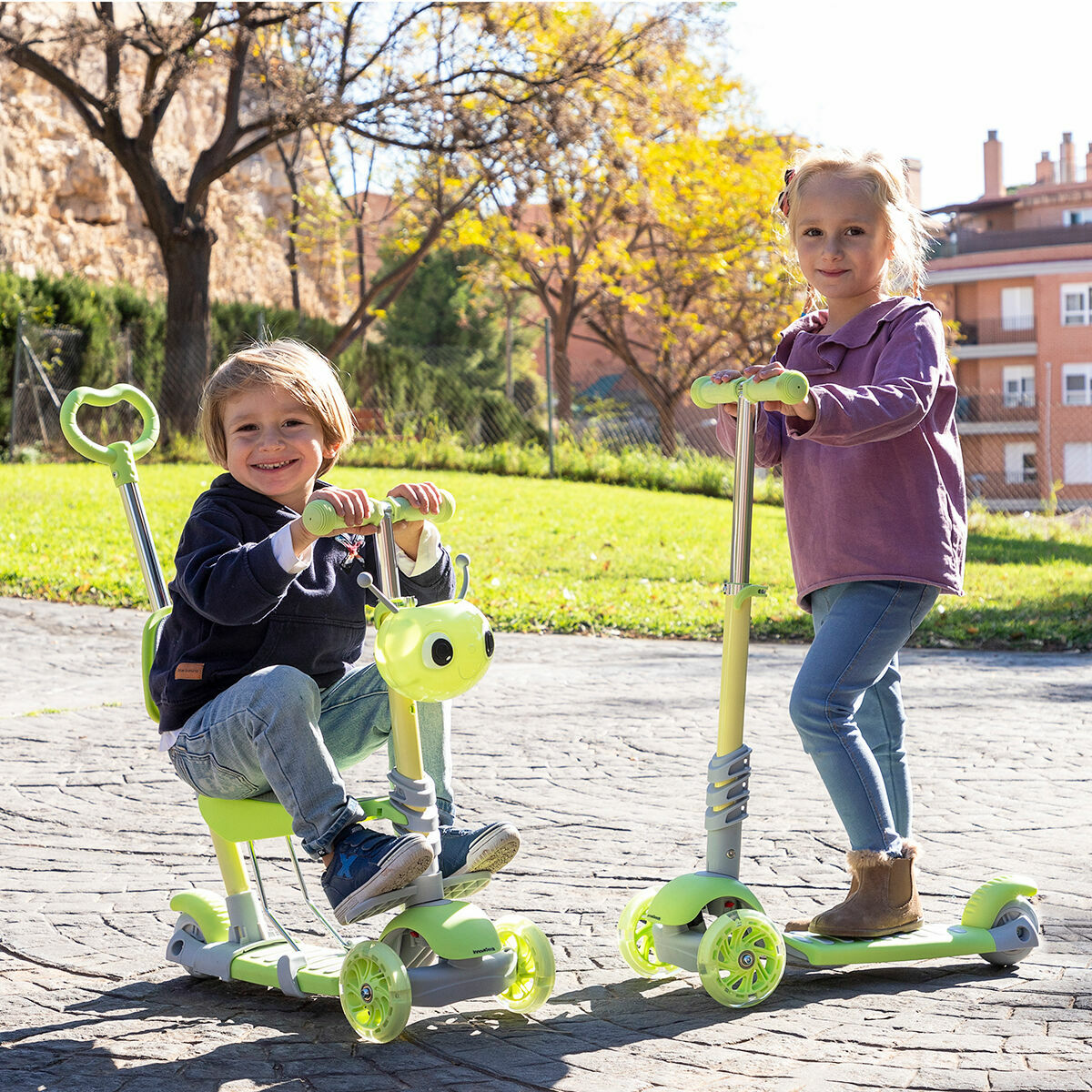 Dos niños montando patinetes, uno sentado, otro de pie, sonriendo, al aire libre.