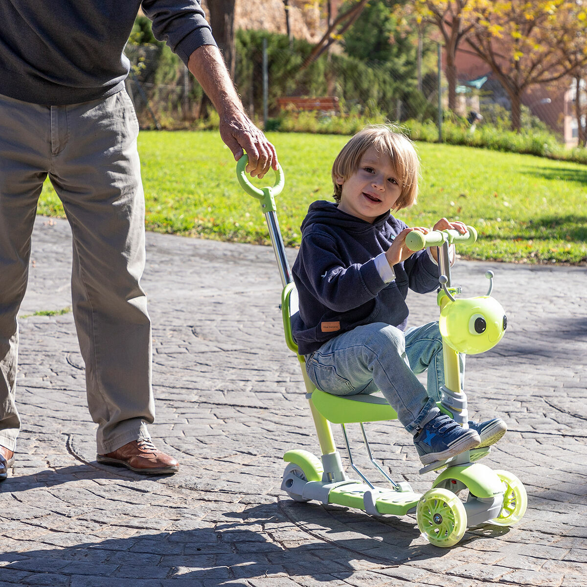 Un niño monta un scooter verde, empujado por un adulto en un parque.