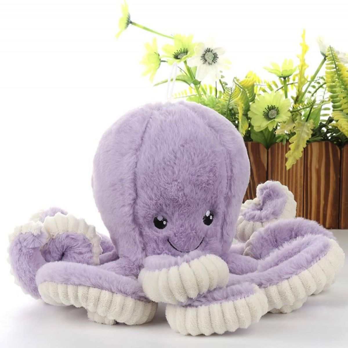 Lila Plüschoktopus mit weißen Akzenten, sitzt auf weißer Oberfläche. Blumen im Hintergrund.