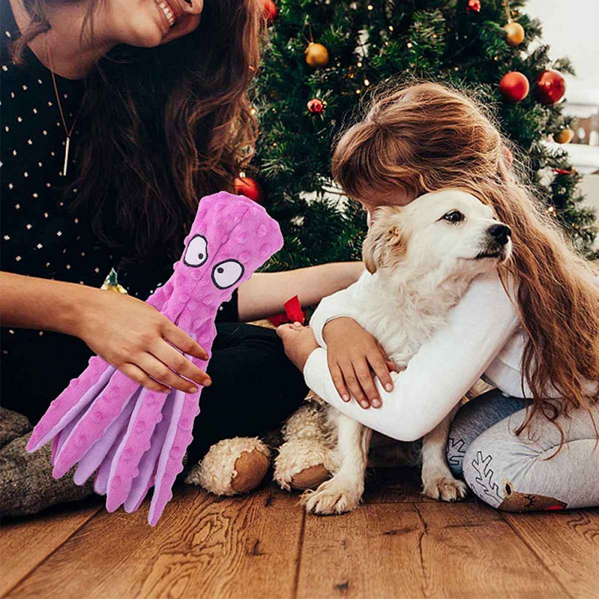 Ein Kind umarmt einen Hund, während eine Frau ein lila Oktopus-Spielzeug hält. Weihnachtsbaum im Hintergrund.