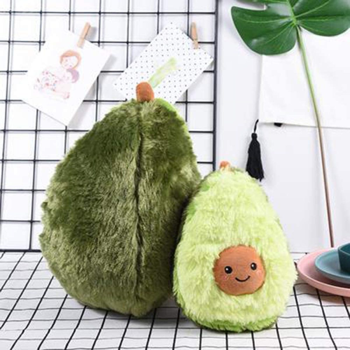 Zwei Plüsch-Avocado-Spielzeuge, eines mit Gesicht, auf weißem Hintergrund, mit einigen dekorativen Gegenständen.