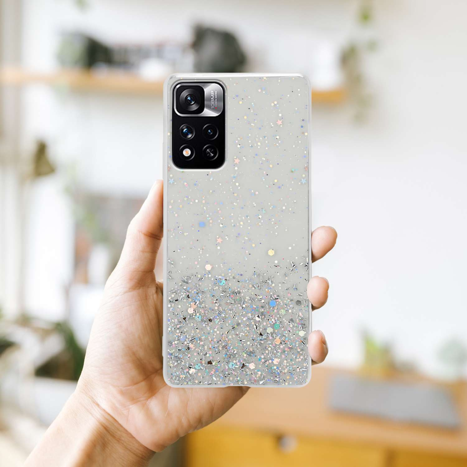 Mano che tiene una custodia trasparente coperta di glitter con fotocamera e sfondo sfocato.