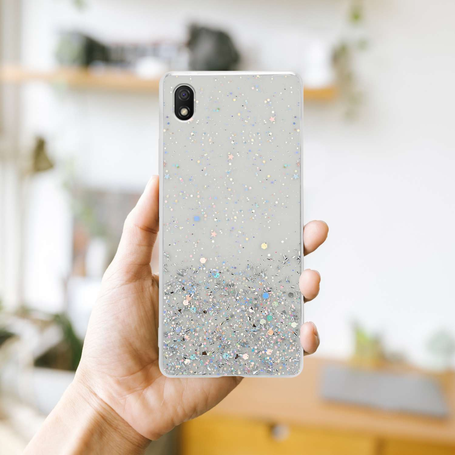 Una mano che tiene un telefono in una custodia trasparente con glitter, su sfondo sfocato.