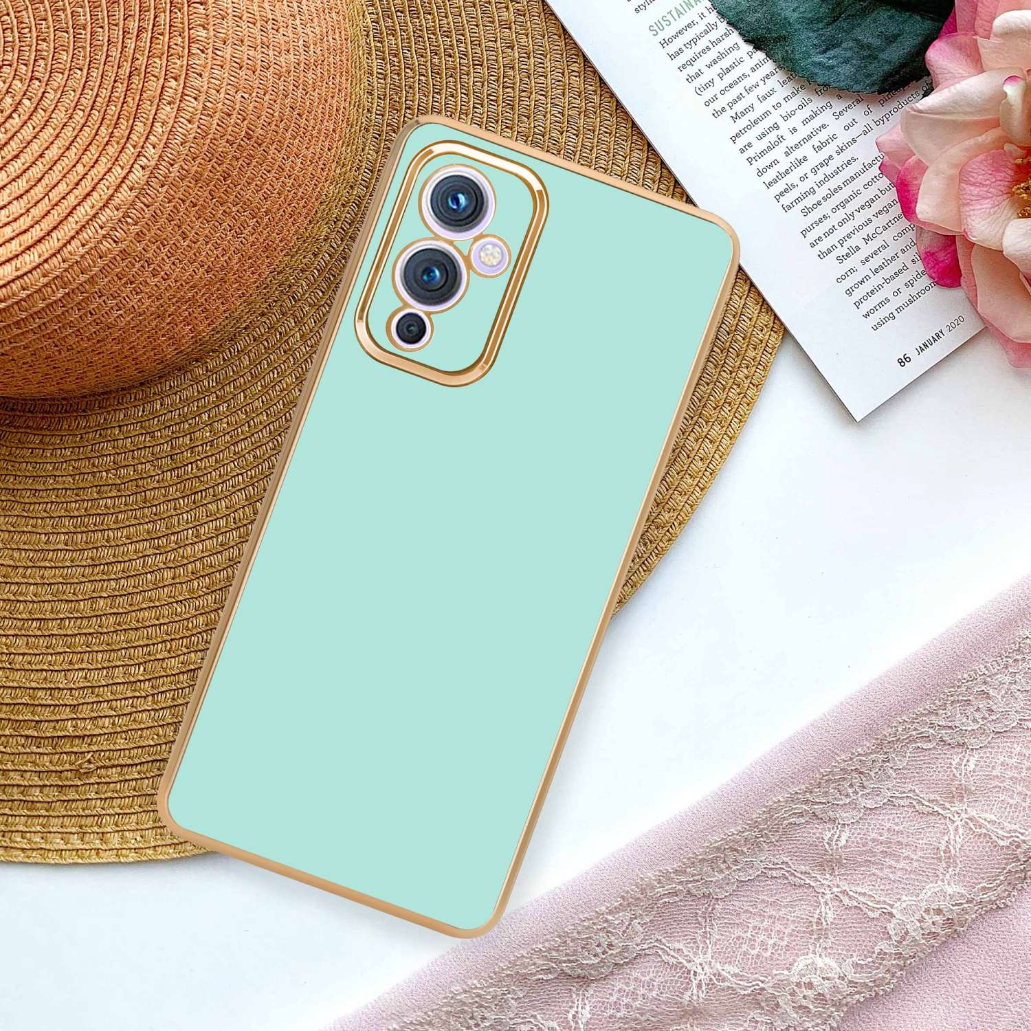 Custodia per telefono verde con finiture dorate, appoggiata su una superficie con un cappello e una sciarpa rosa.