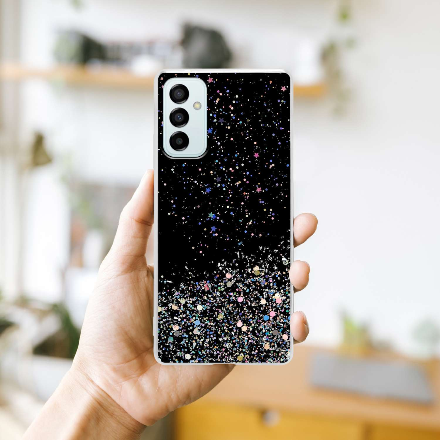 Mano che tiene custodia nera con glitter, mostrando telefono con tripla fotocamera.