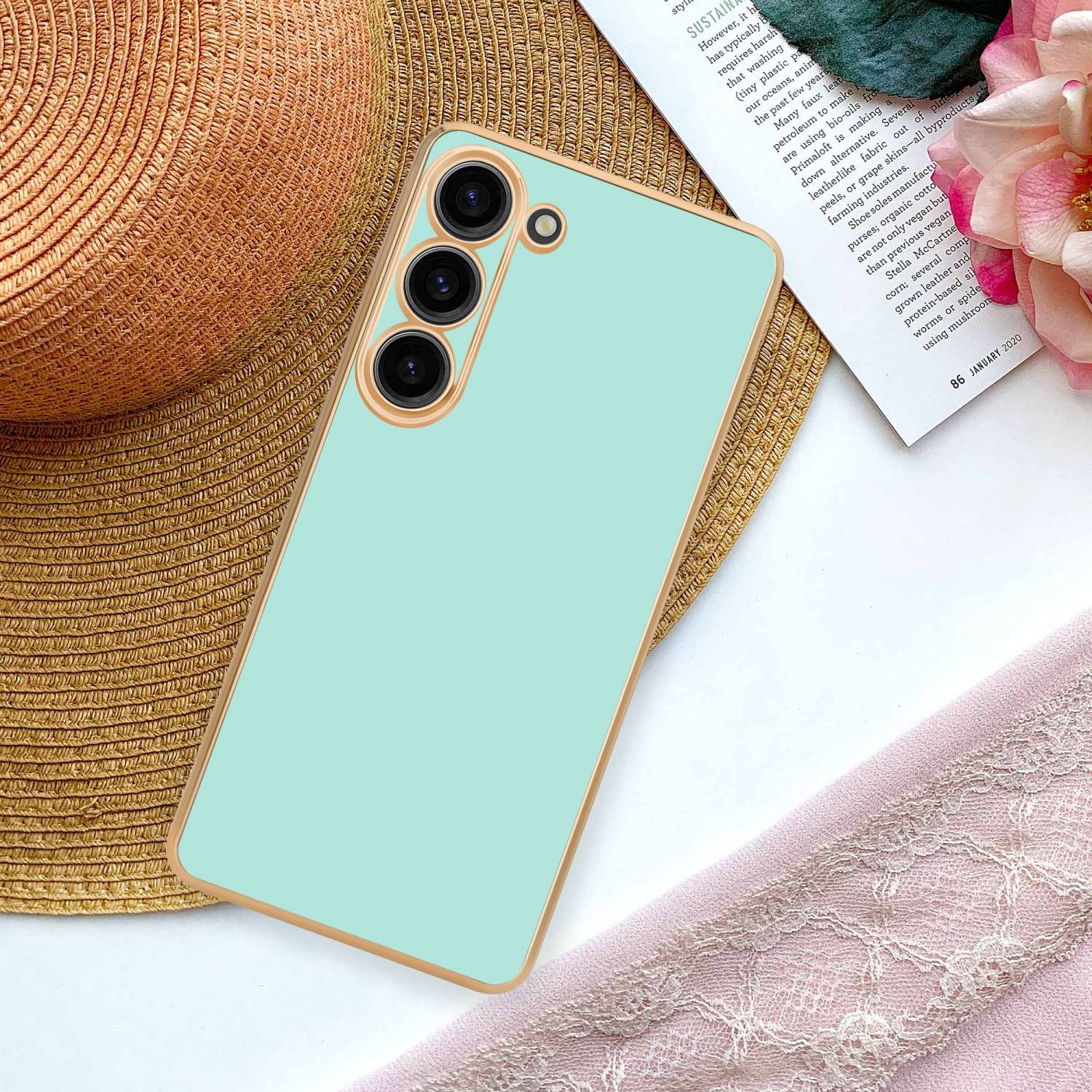 Una custodia per telefono verde chiaro con bordo dorato su un cappello di paglia, accanto a una sciarpa rosa e un fiore.