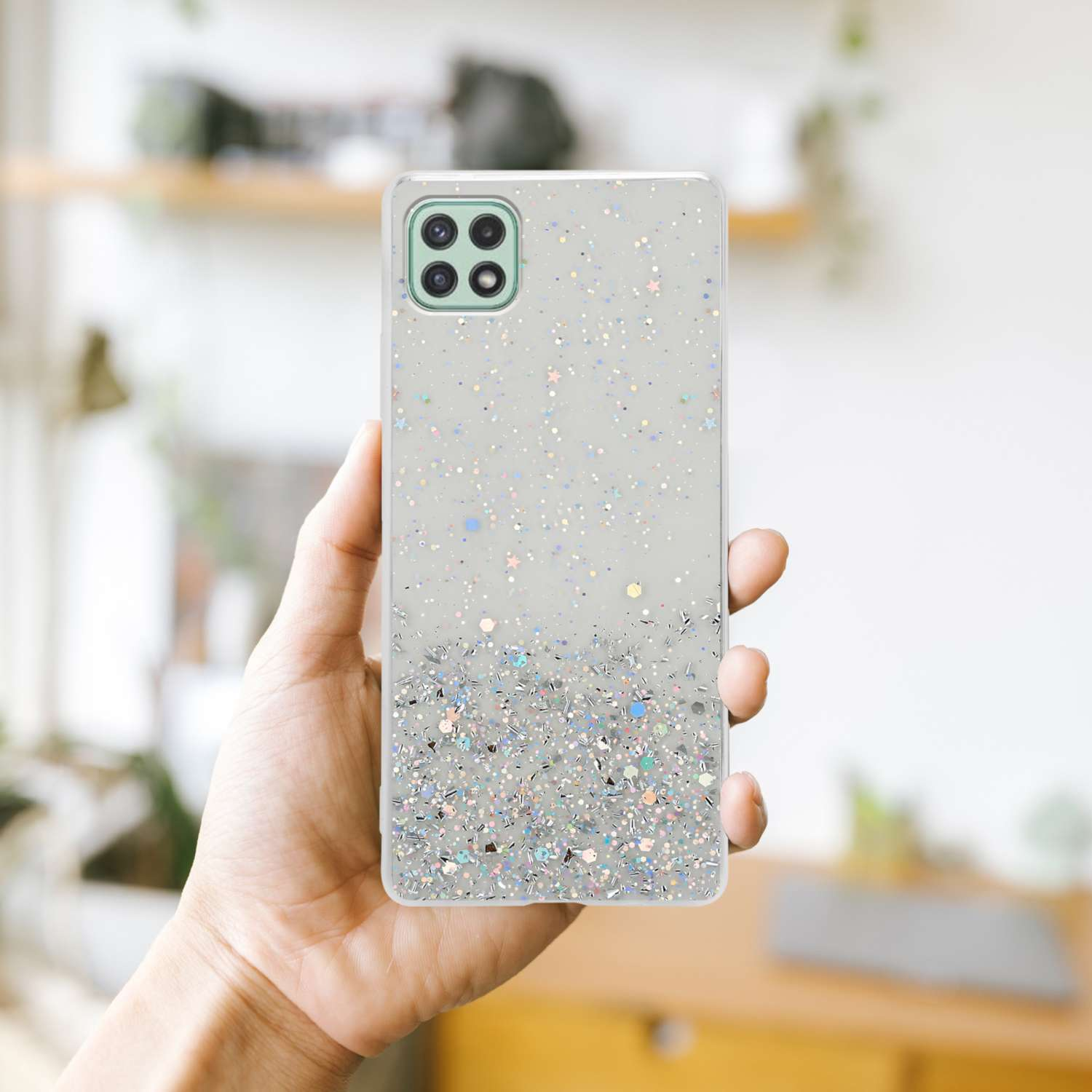 Mano che tiene un telefono con custodia glitterata, con sfondo sfocato, al chiuso.