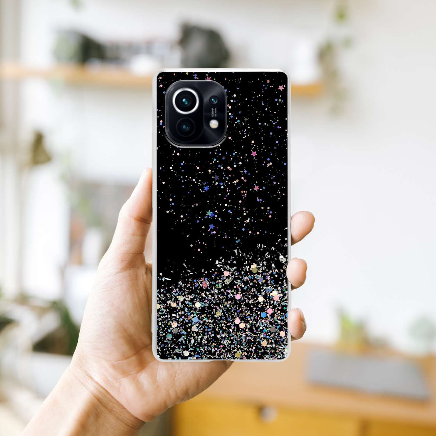 Mano che tiene un telefono in una custodia nera, con un design glitterato su uno sfondo sfocato di una stanza.