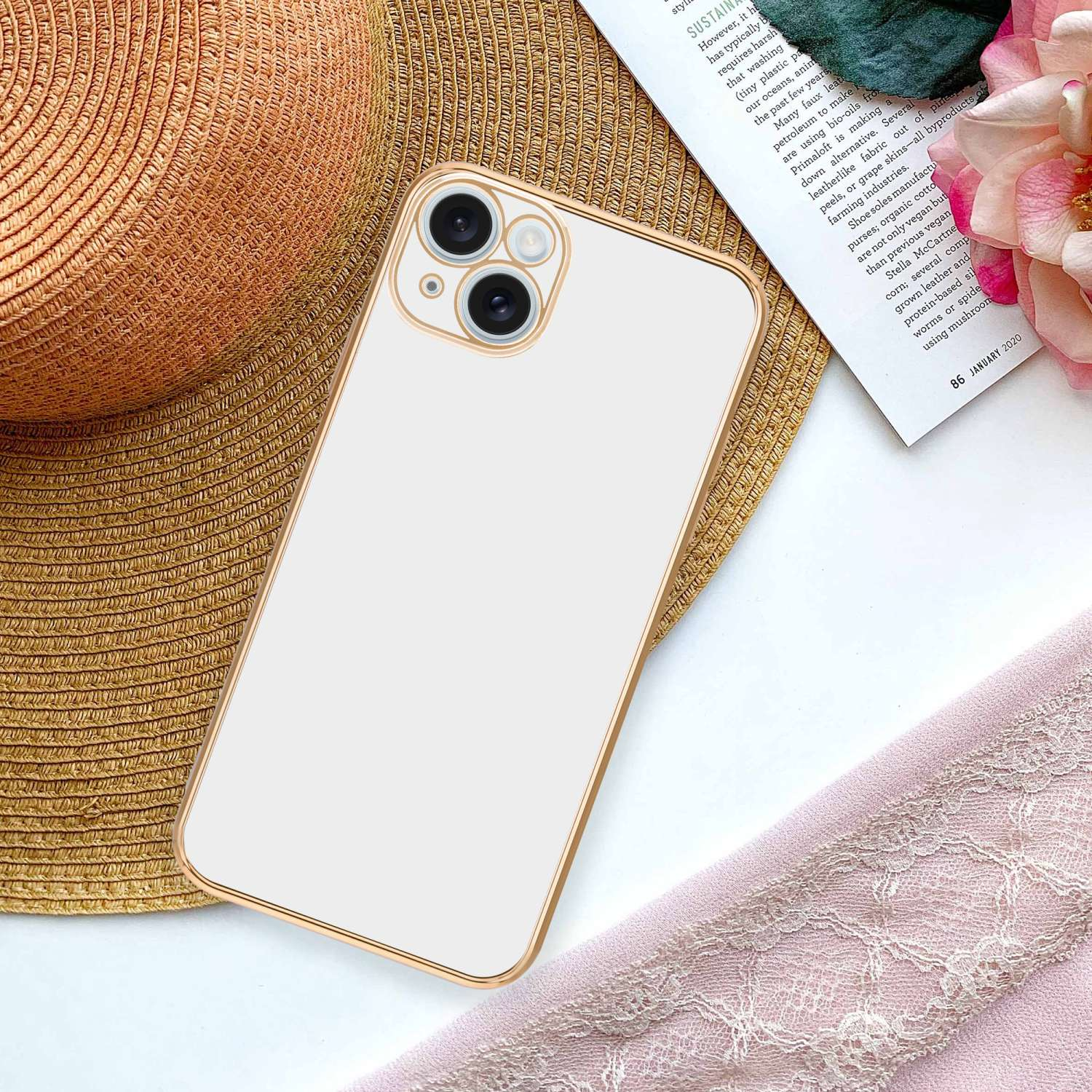 Weißes Telefon mit goldenem Rahmen auf einem Magazin und rosa Stoff, mit Strohhut im Hintergrund.
