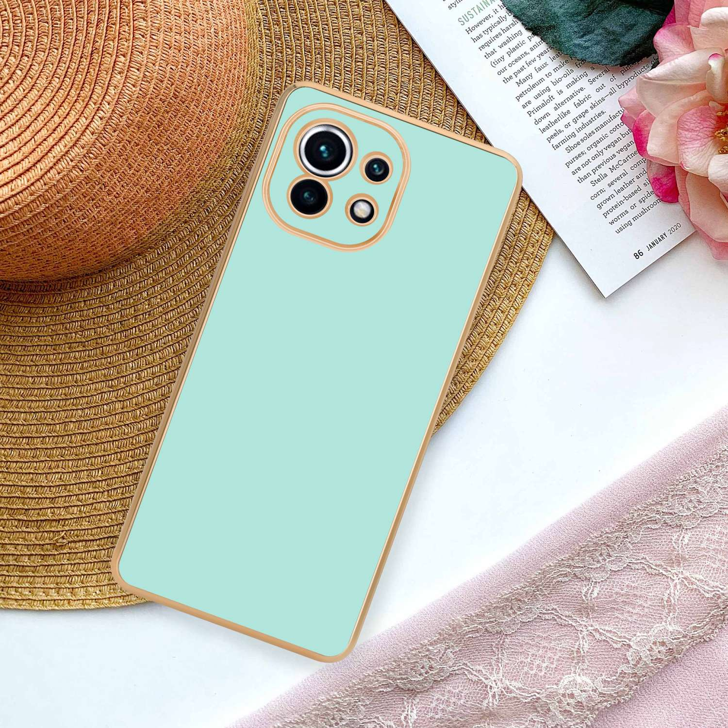 Custodia per telefono turchese con finiture dorate su cappello di paglia, accanto a rivista e fiore rosa.