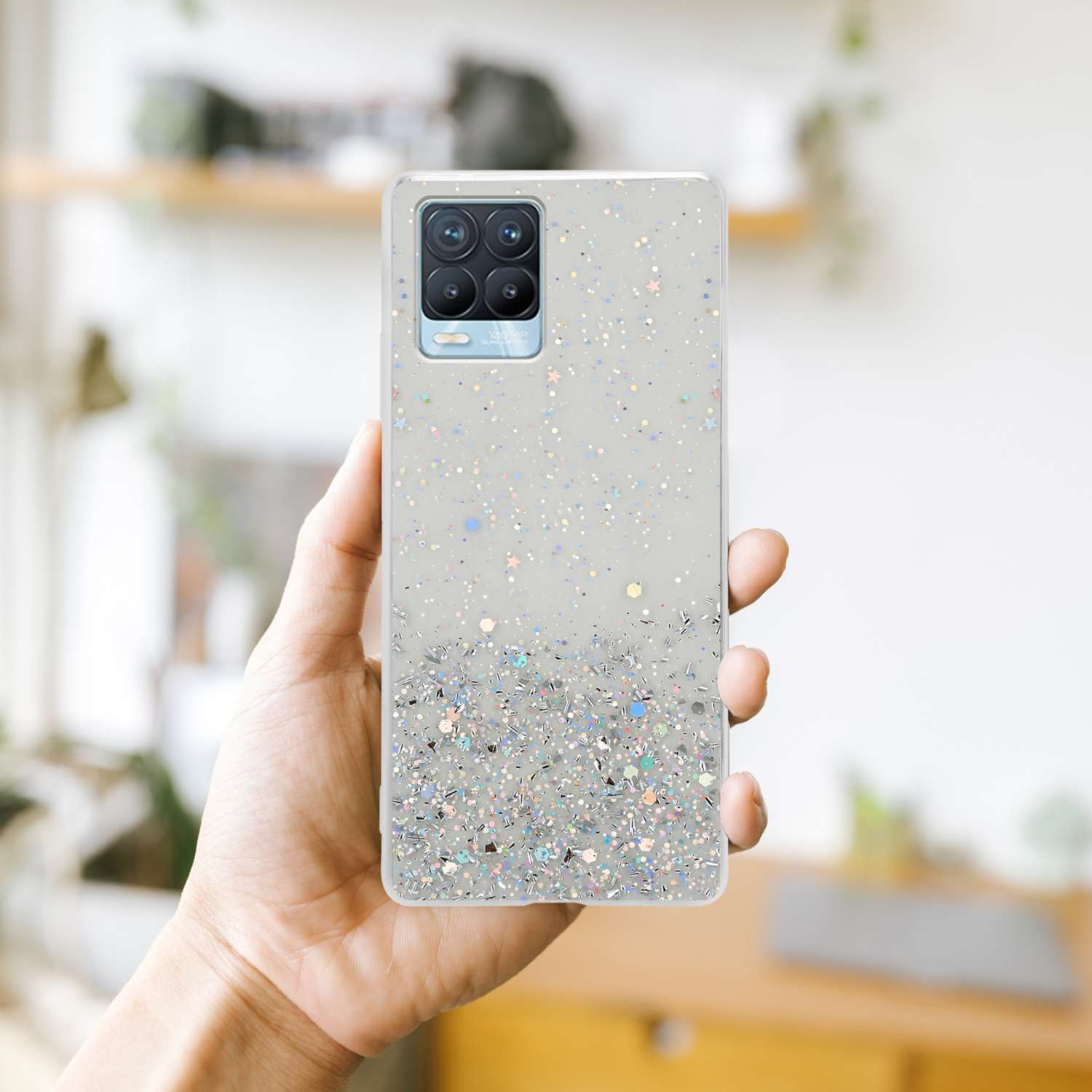 Mano che tiene una custodia per telefono con glitter argentati. La custodia copre il retro di un telefono. Sfondo sfocato.