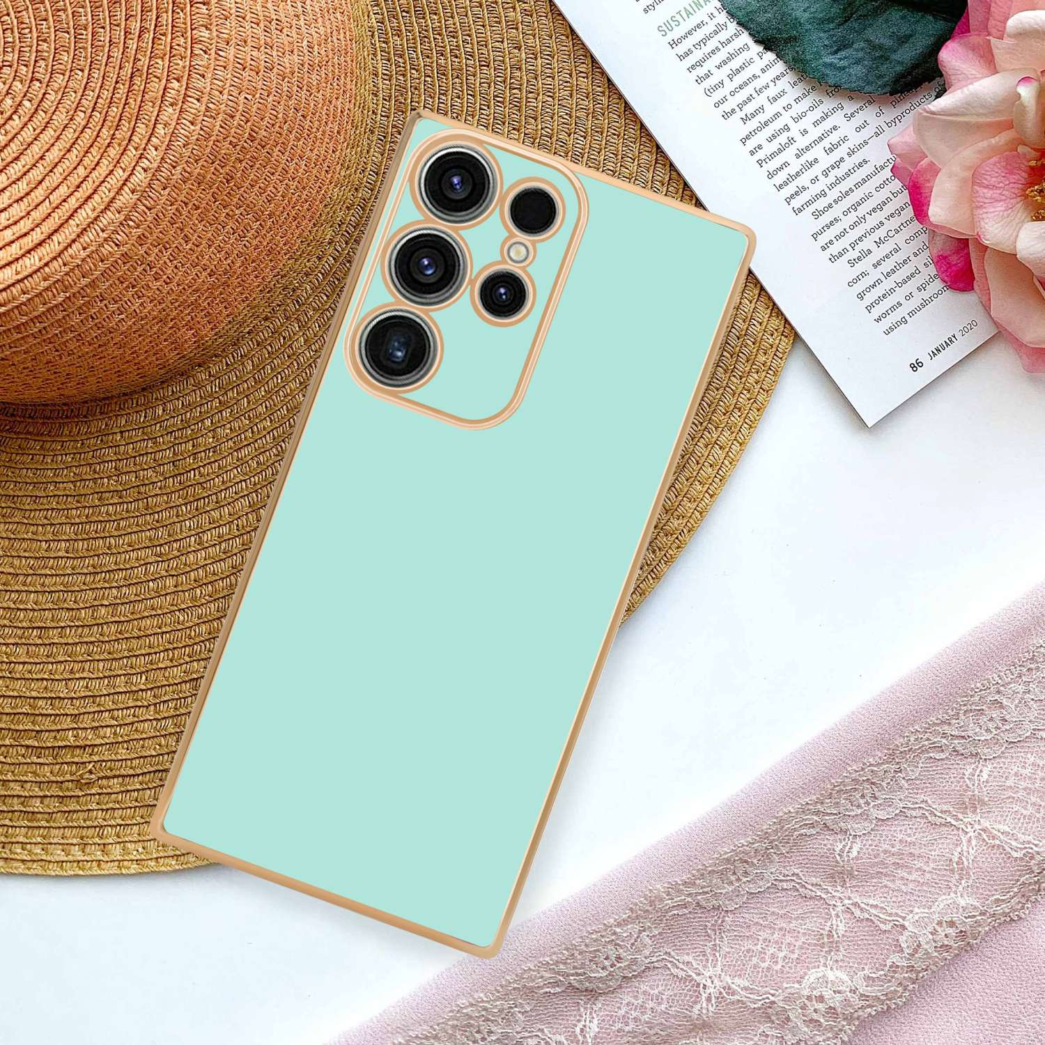 Custodia per telefono verde menta con finiture dorate, appoggiata su un panno rosa, con un cappello.