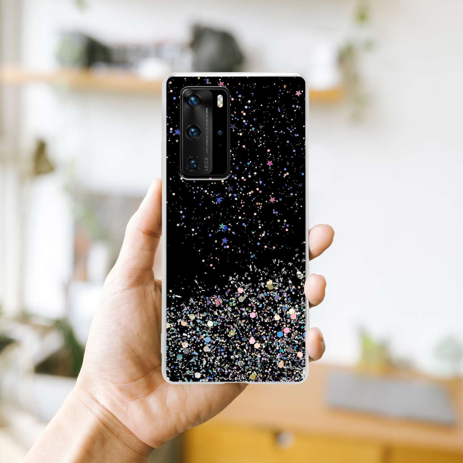 Eine Hand hält ein Telefon mit schwarzem und Glitzerdesign. Der Hintergrund ist ein verschwommenes Raumambiente.