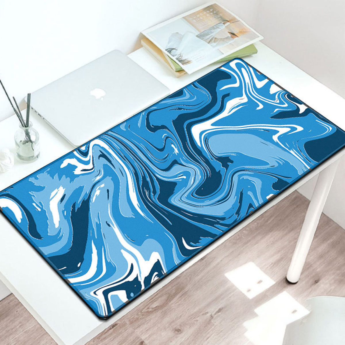Blau-weiß abstrakt gestaltetes Mauspadd auf einem weißen Schreibtisch. Laptop und Stifte sind in der Nähe.