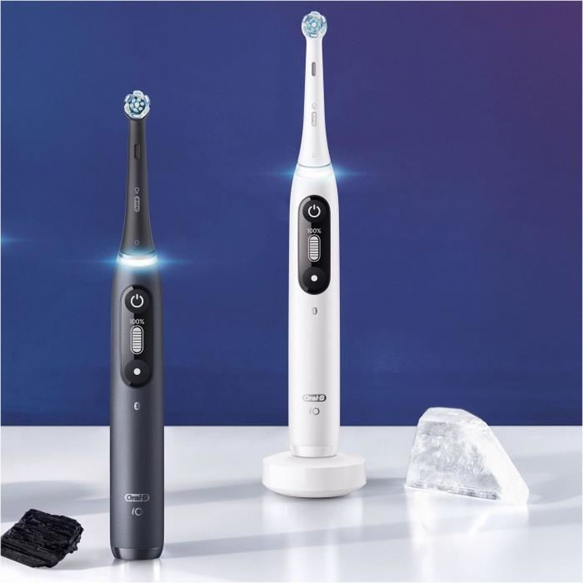Dos cepillos de dientes eléctricos. Uno negro, uno blanco, ambos con luces encendidas. Una base de carga y un cristal en el fondo.
