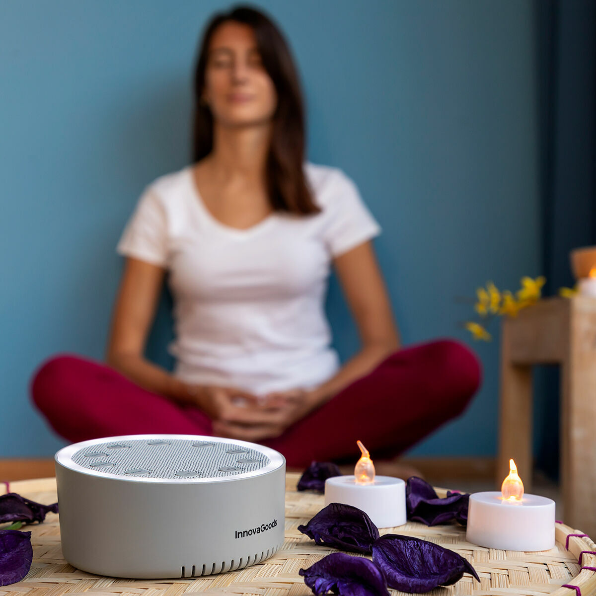 Mujer meditando con máquina de sonido, velas y pétalos morados sobre esterilla tejida. Pared azul.