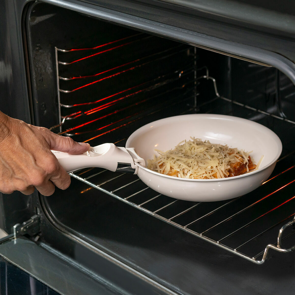 Una mano rimuove una padella bianca con cibo da un forno nero. Il cibo è ricoperto di formaggio grattugiato.