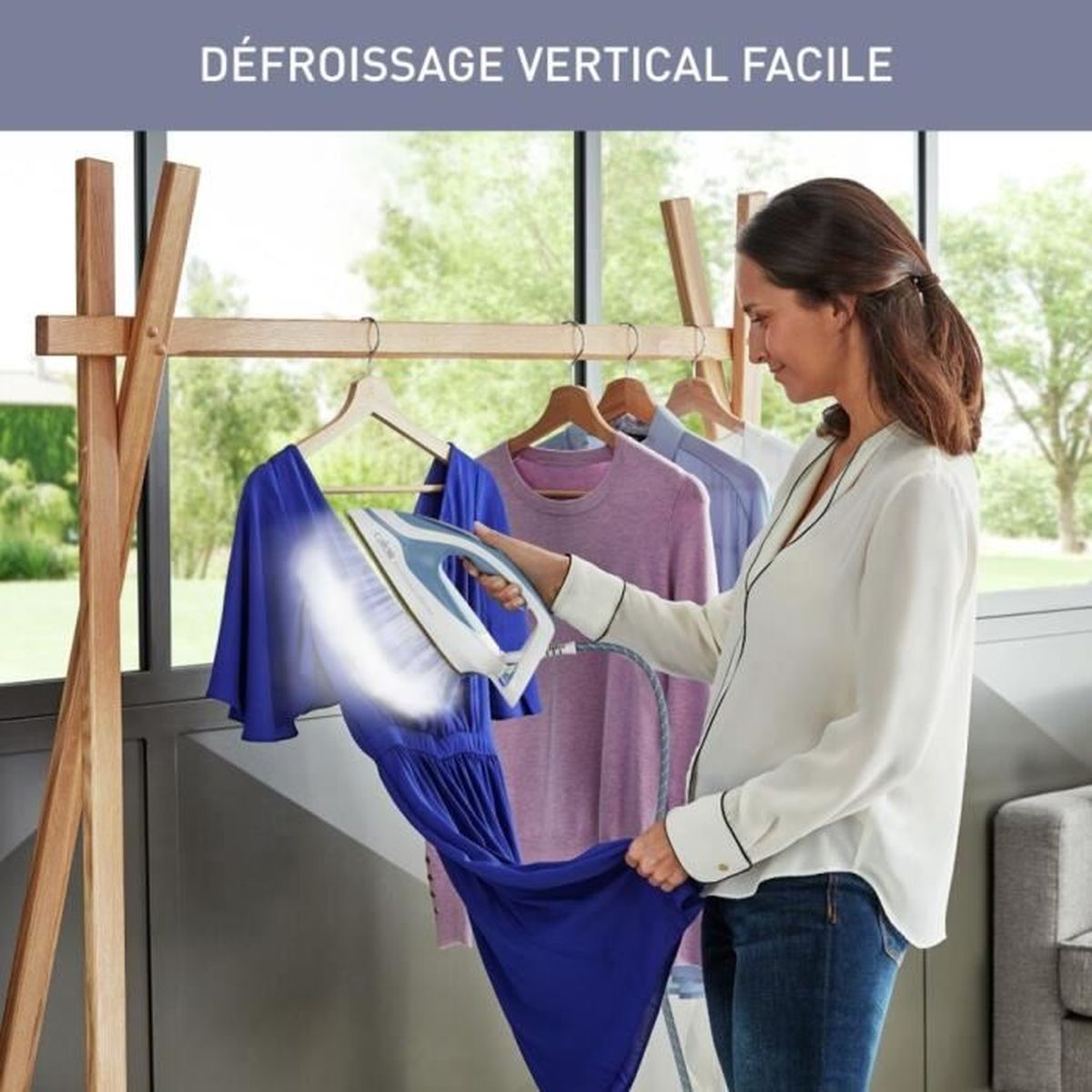 Donna che stira un vestito blu a vapore. Un appendiabiti in legno contiene vestiti, finestra sullo sfondo.