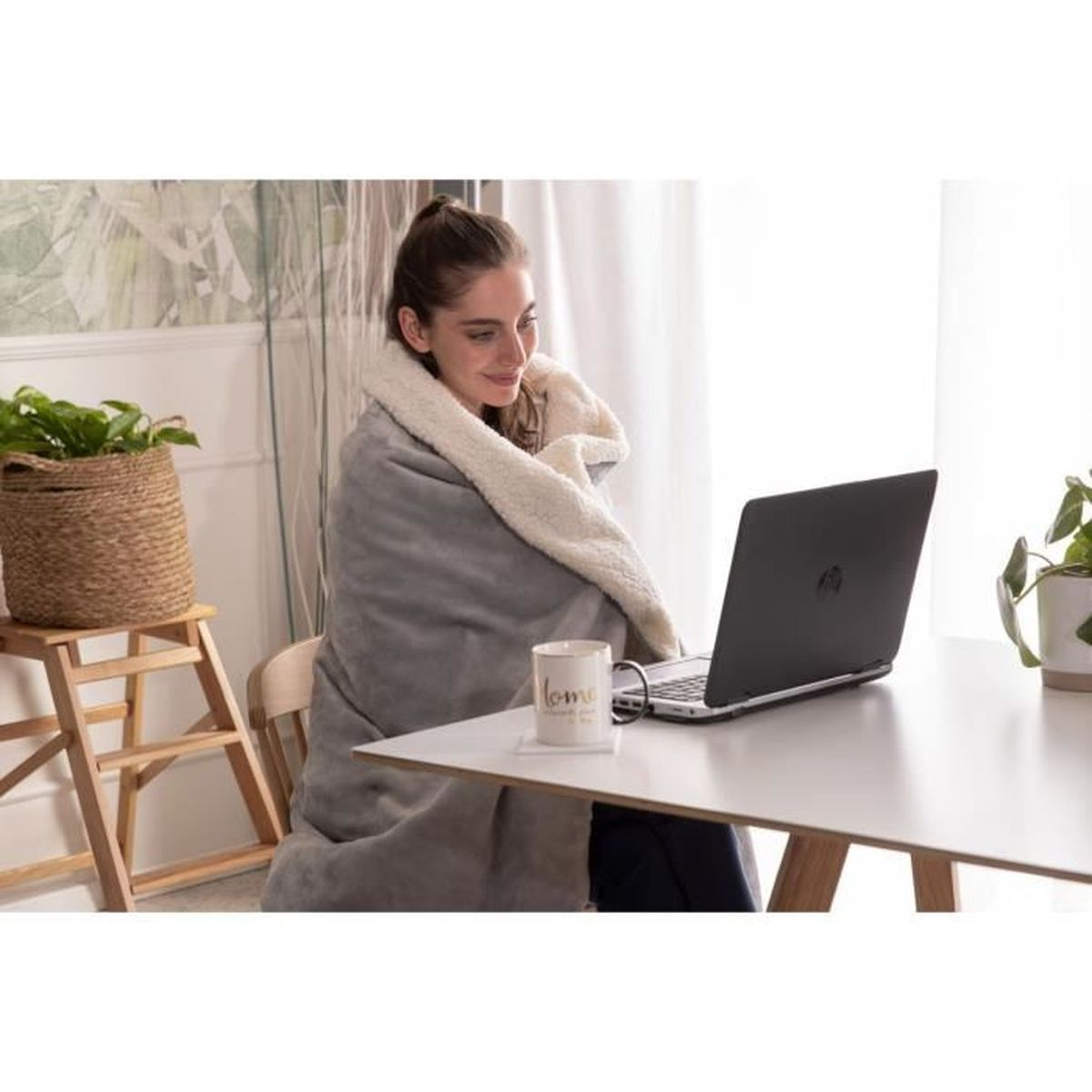 Una donna è seduta su una sedia con una coperta e lavora su un laptop. C'è una tazza sul tavolo.