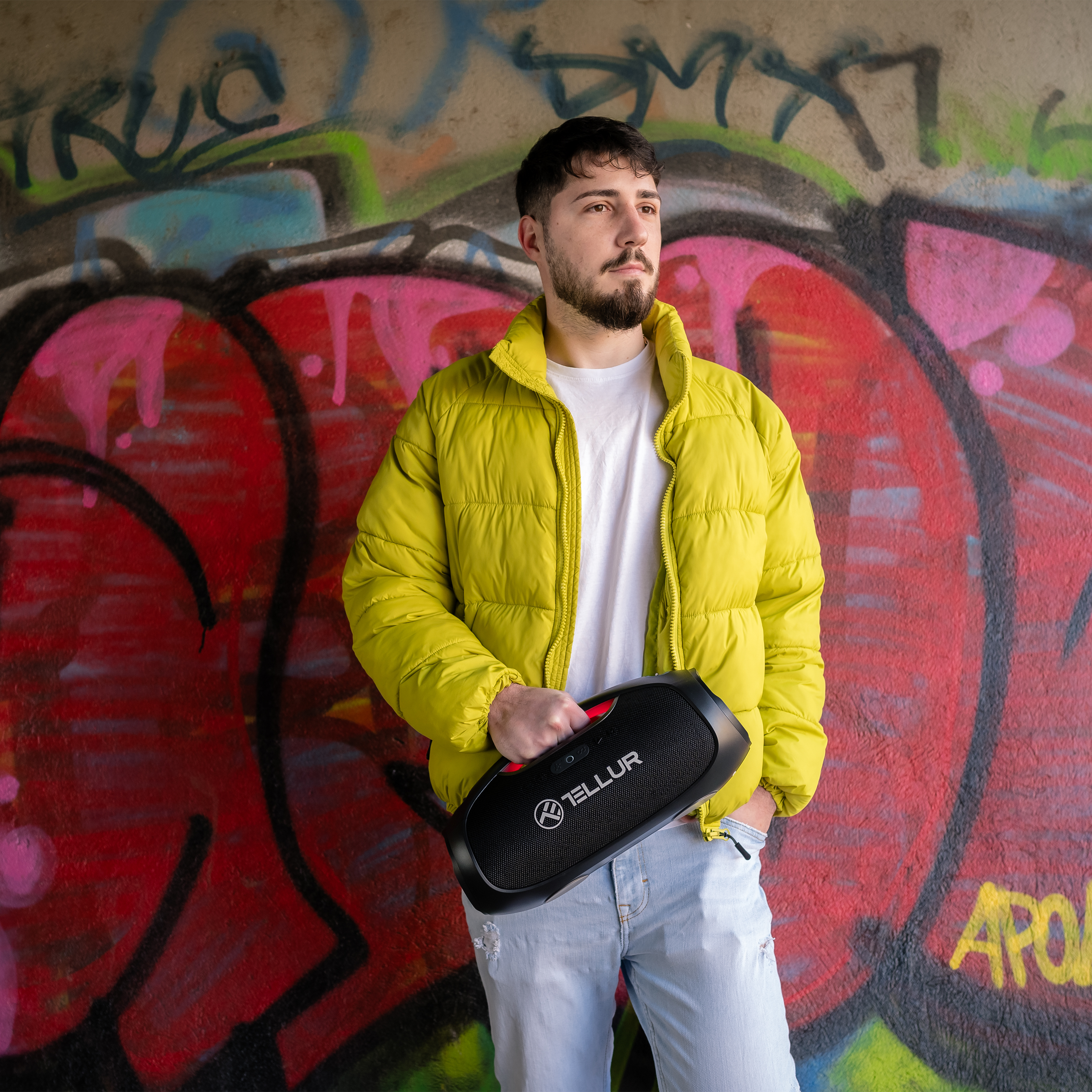 Mężczyzna trzymający czarny głośnik. Ma na sobie żółtą kurtkę przed graffiti.