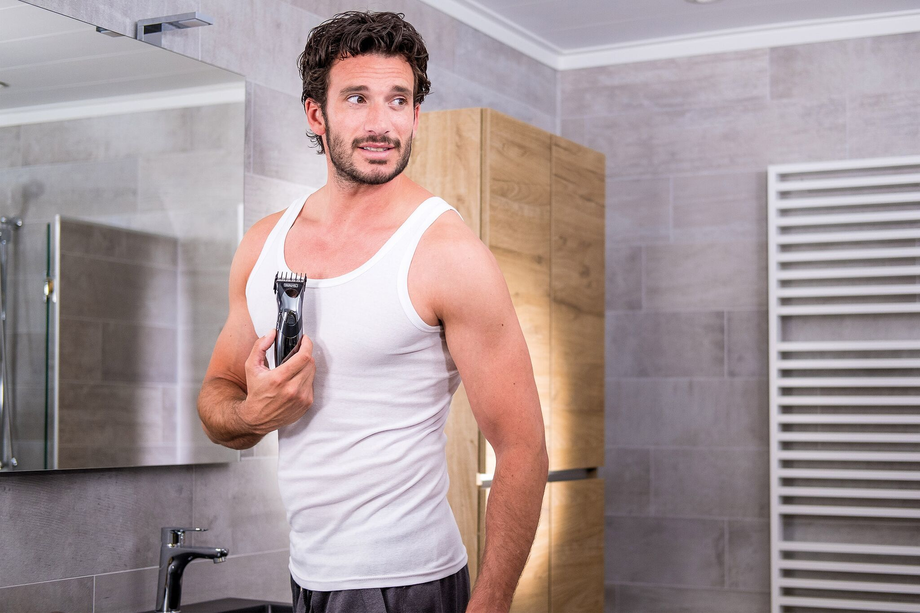 Mann rasiert Brusthaare mit Trimmer im Badezimmer. Spiegel und Schränke im Hintergrund.