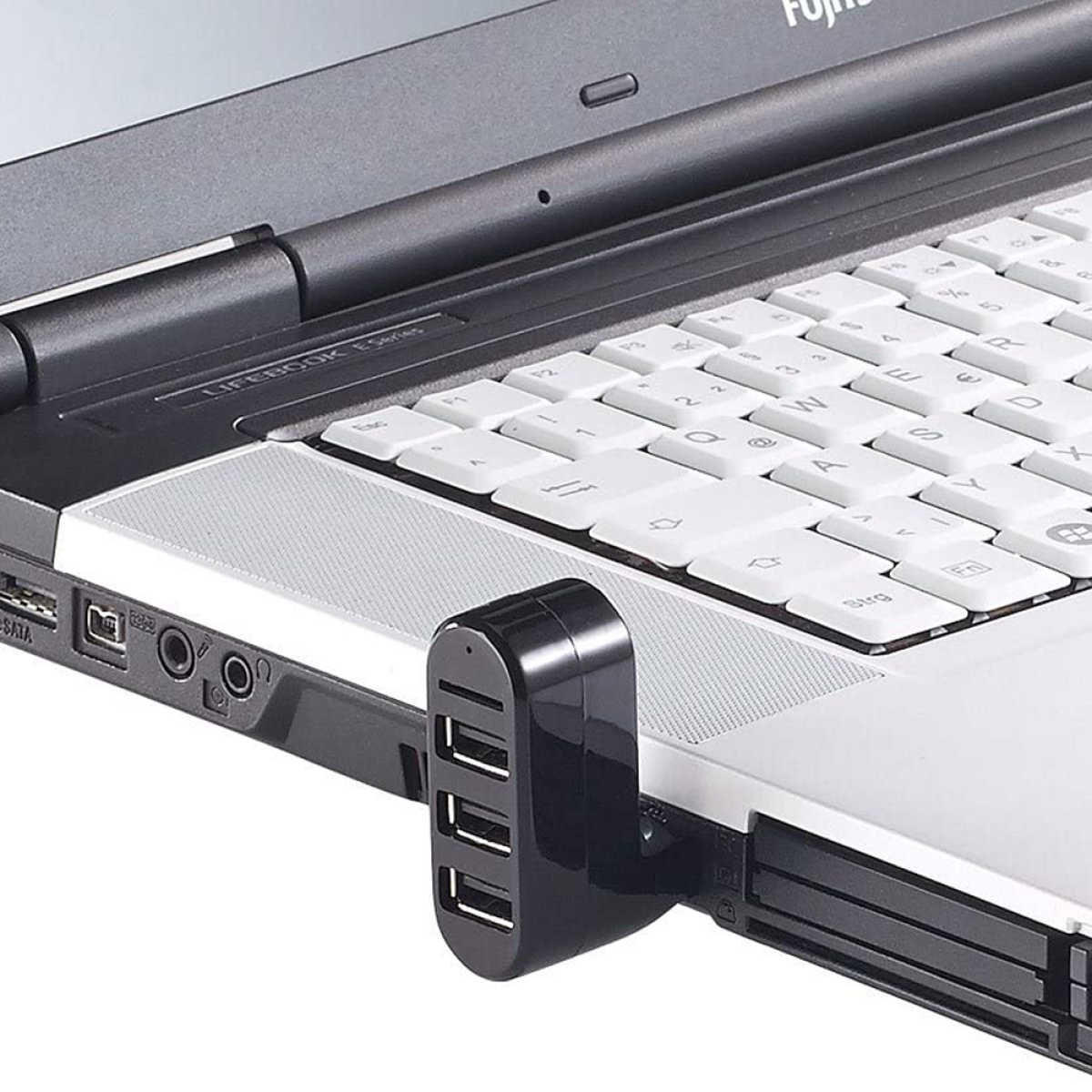 Ein Laptop ist geöffnet, mit einem USB-Hub an der Seite befestigt. Der Laptop steht auf einer weißen Oberfläche.