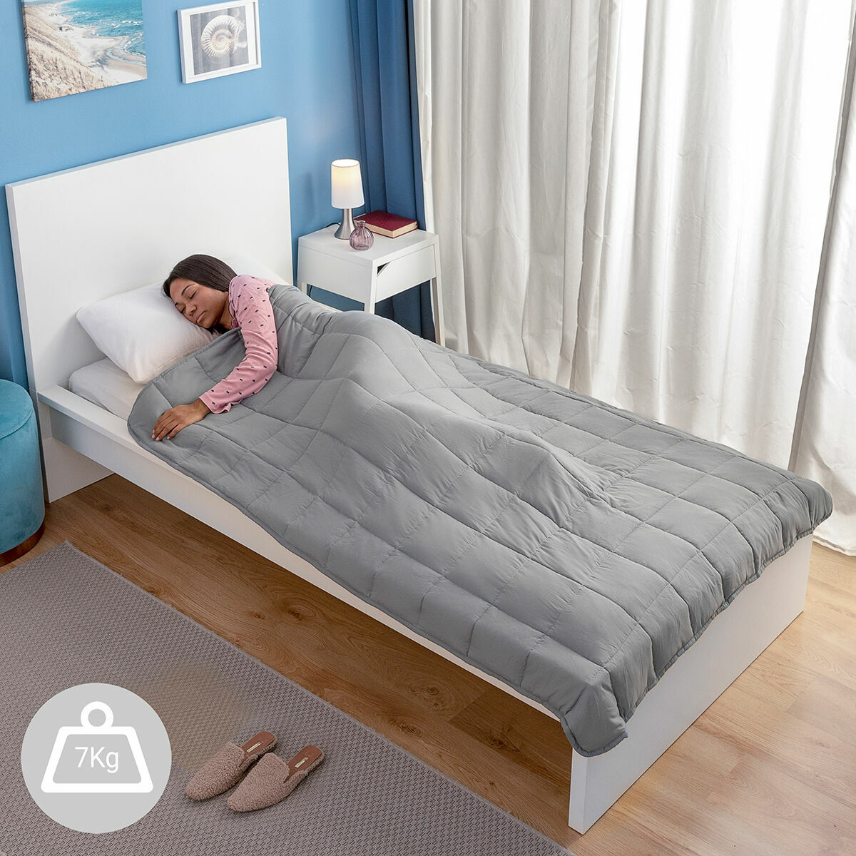 Una donna dorme su un letto coperto da una coperta grigia ponderata. Etichetta del peso da 7 kg visibile. Stanza con mobili.