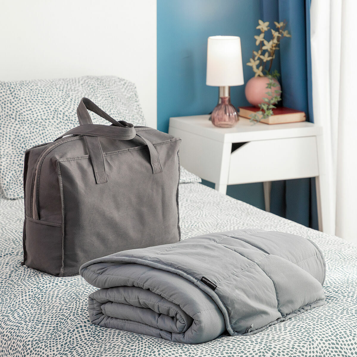 Una coperta grigia ponderata e una borsa abbinata sono su un letto. Un comodino e una lampada sono visibili.
