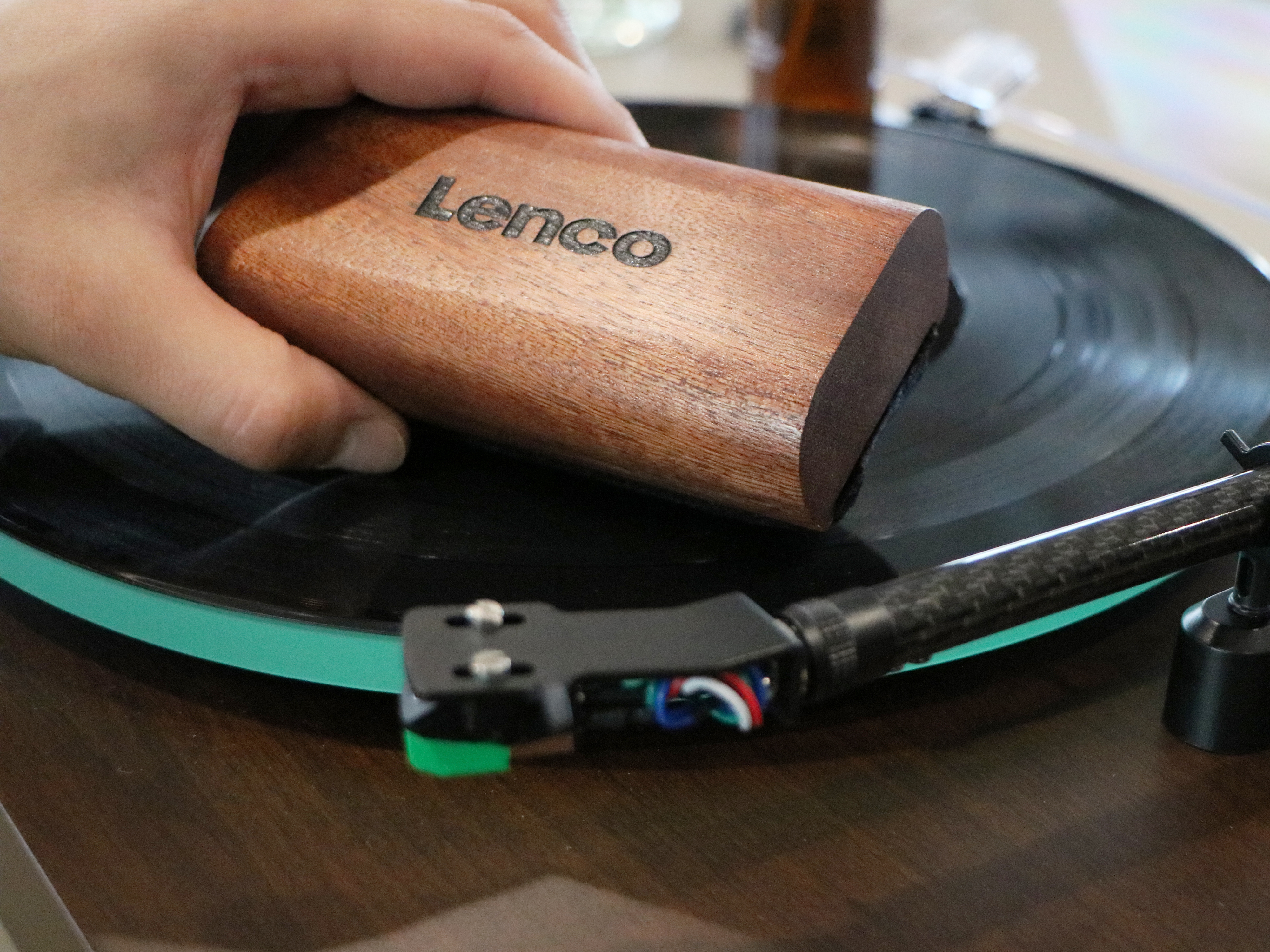 Een hand die een vinylplaat schoonmaakt met een houten Lenco-borstel, draaitafel op de achtergrond.