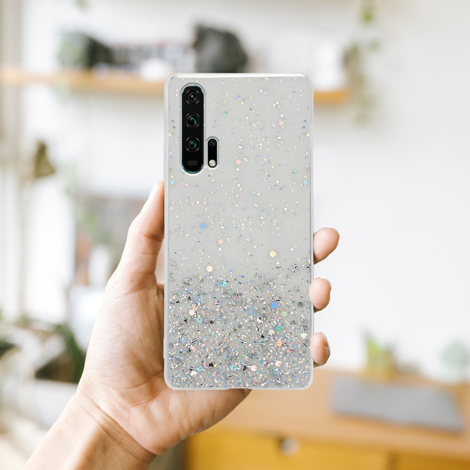 Una mano tiene un telefono con una custodia glitterata. Lo sfondo è un muro bianco sfocato con qualche decorazione.