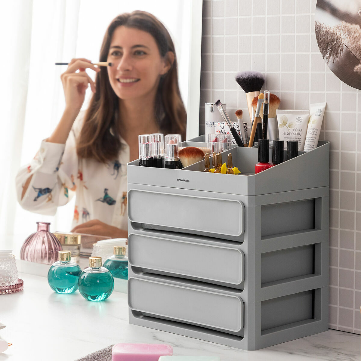 Frau schminkt sich an einer Kommode. Make-up und Pinsel sind in einem grauen, mehrstufigen Organizer.