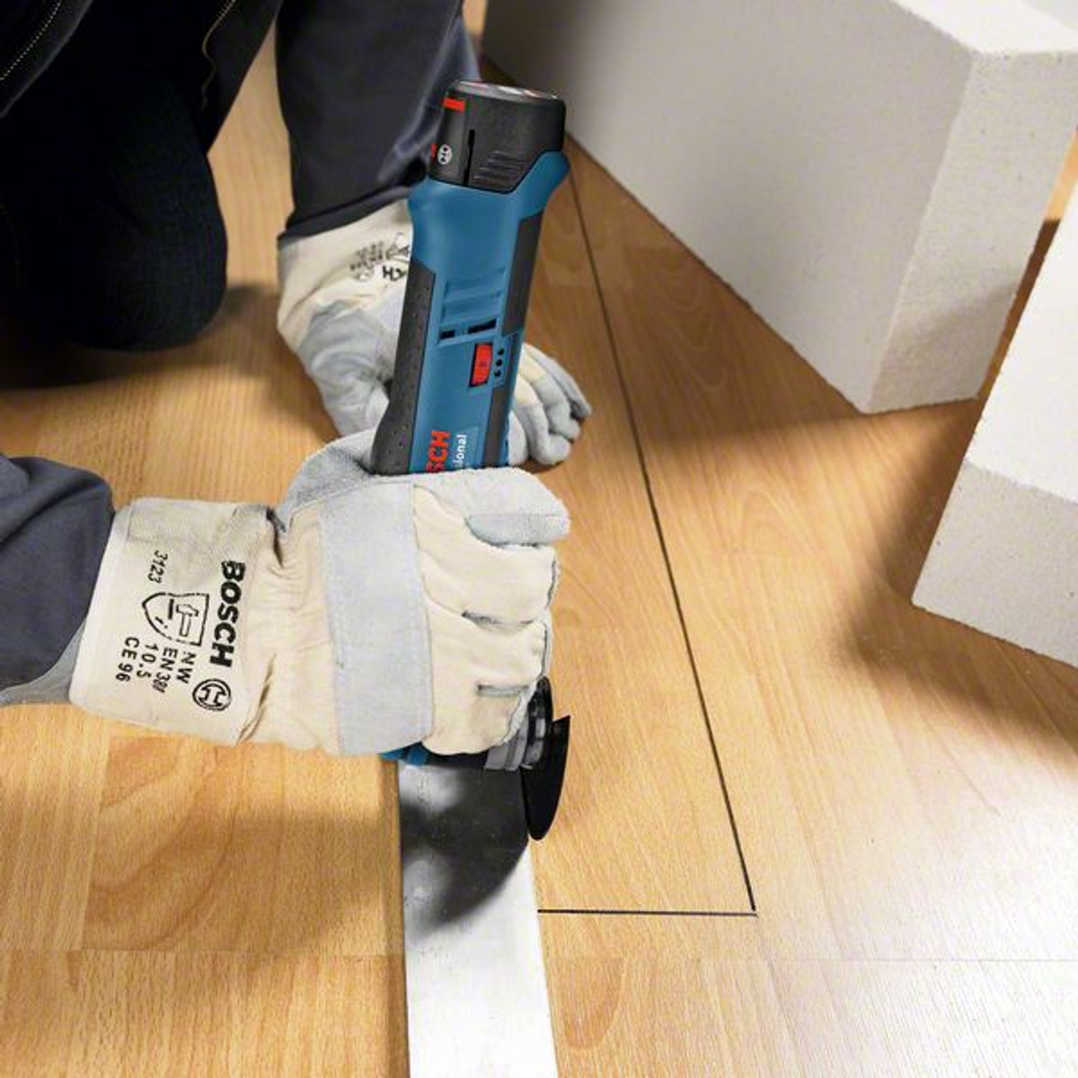 Eine Person mit Handschuhen schneidet mit einer Bosch-Säge eine Linie in einen Holzboden. Weiße Schaumstoffblöcke im Hintergrund.