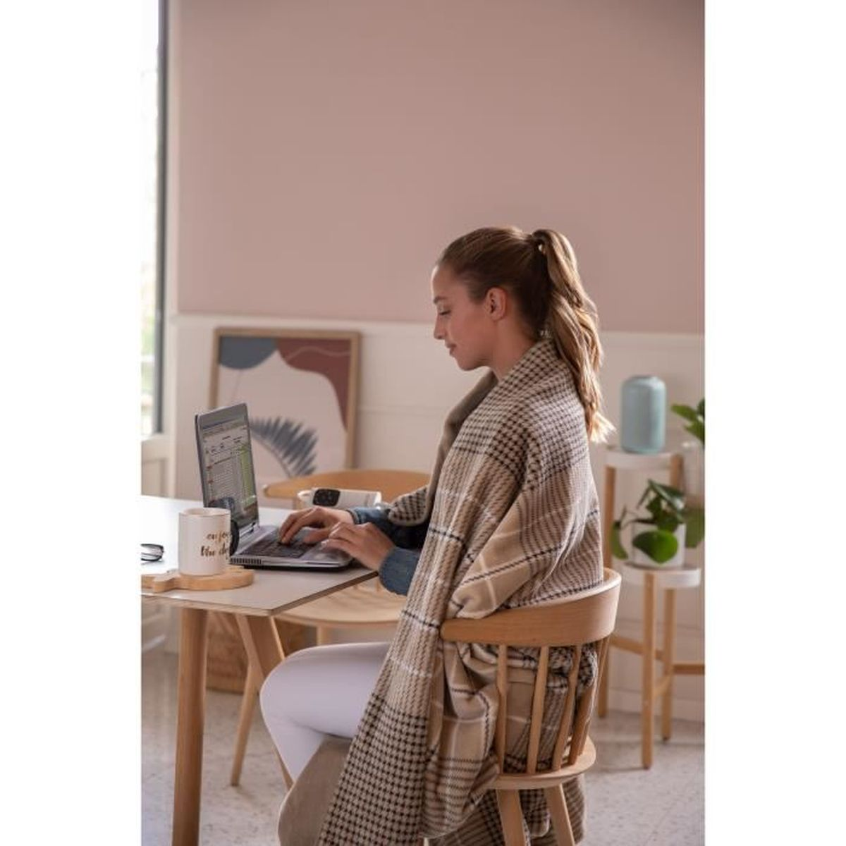 Kobieta korzystająca z laptopa przy stole, w szalu, z kubkiem i lampą.