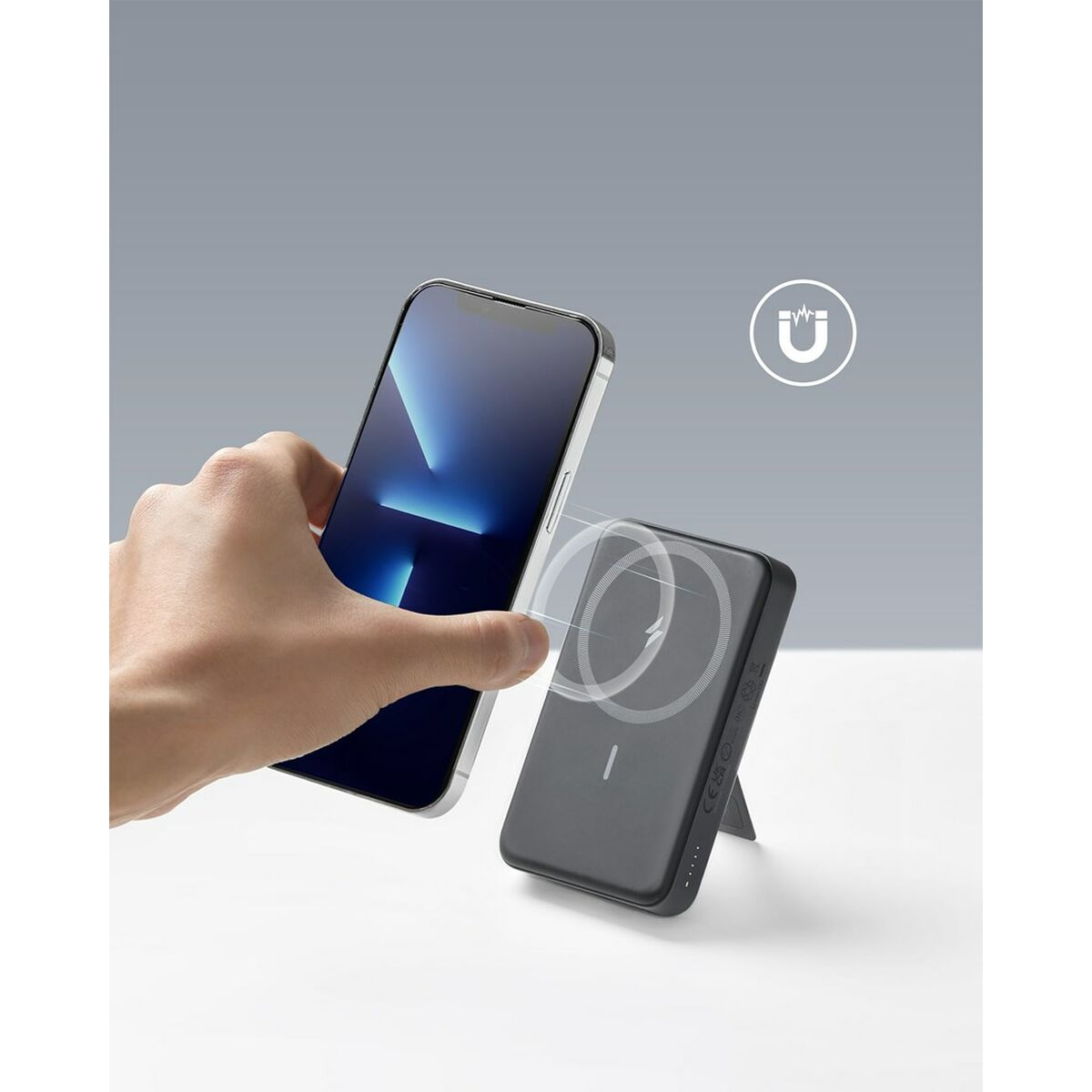 Eine Hand, die ein Telefon neben einem Ladegerät hält. Das Telefon ist schwarz. Grauer Hintergrund.
